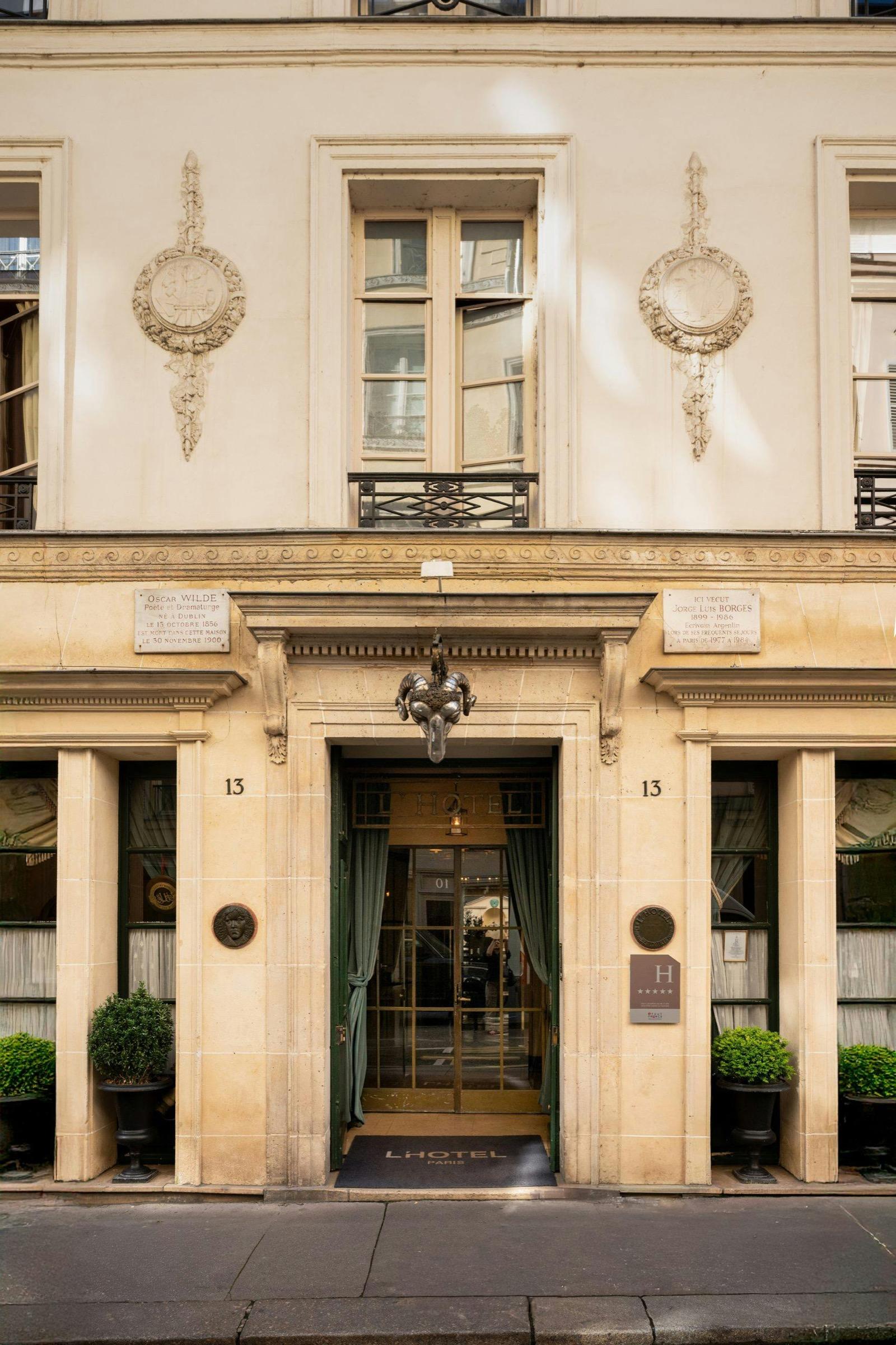 The hotel's cream stone facade and glass entrance doors at street level, flanked by potted plants and marked with the number 13 and a doormat reading 'L'Hotel Paris'.