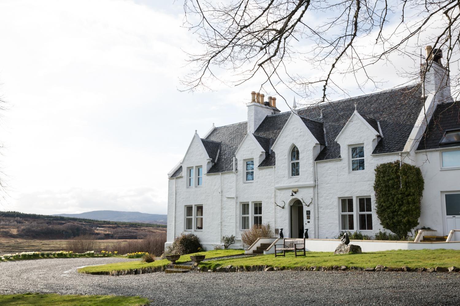 A long two-storey white harled house with grey slate roofs and gabled dormers, seen from the gravel drive with a wooden bench and clipped ivy by the front door.
