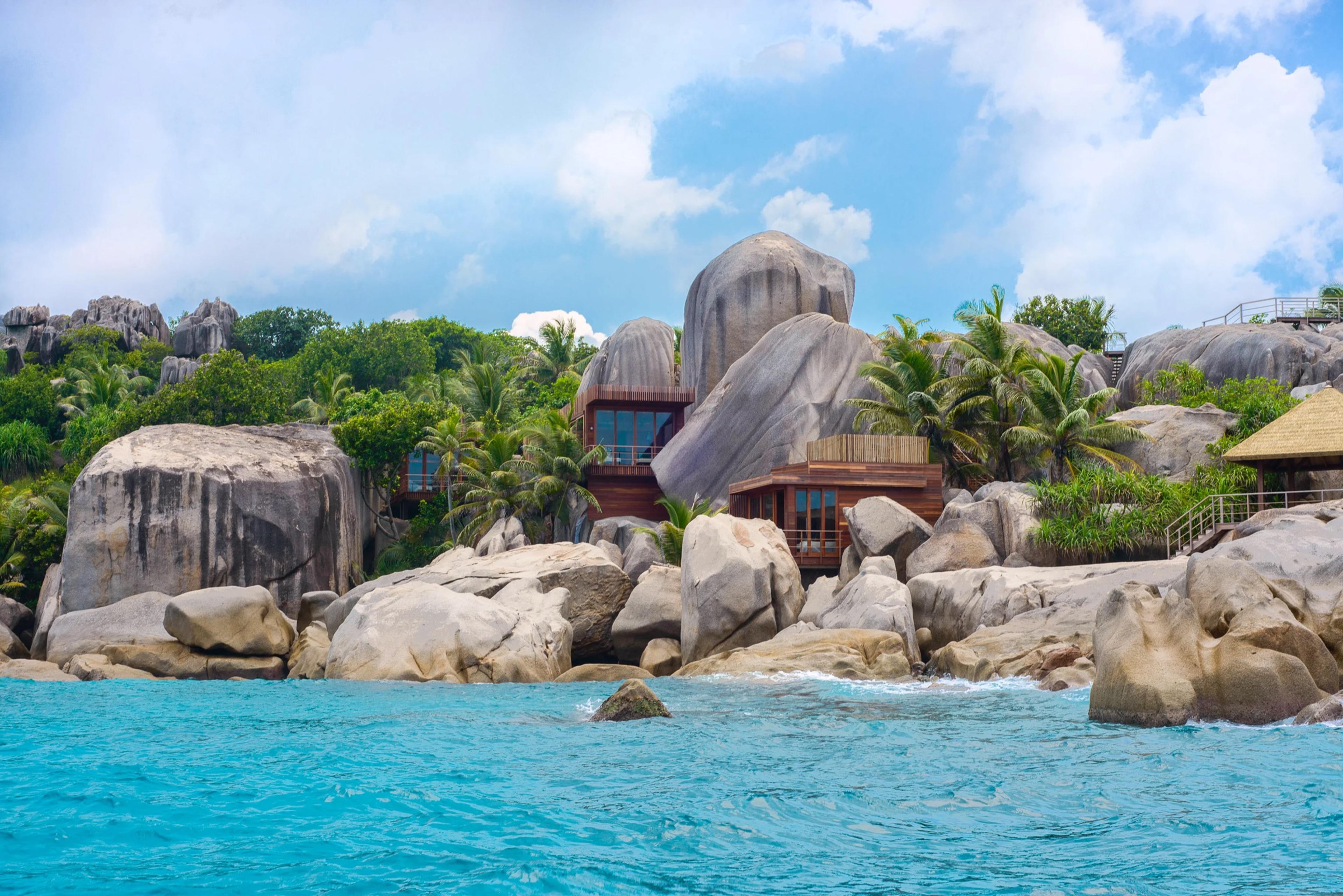 A timber house built among granite boulders and palm trees beside clear turquoise water, seen from the sea.