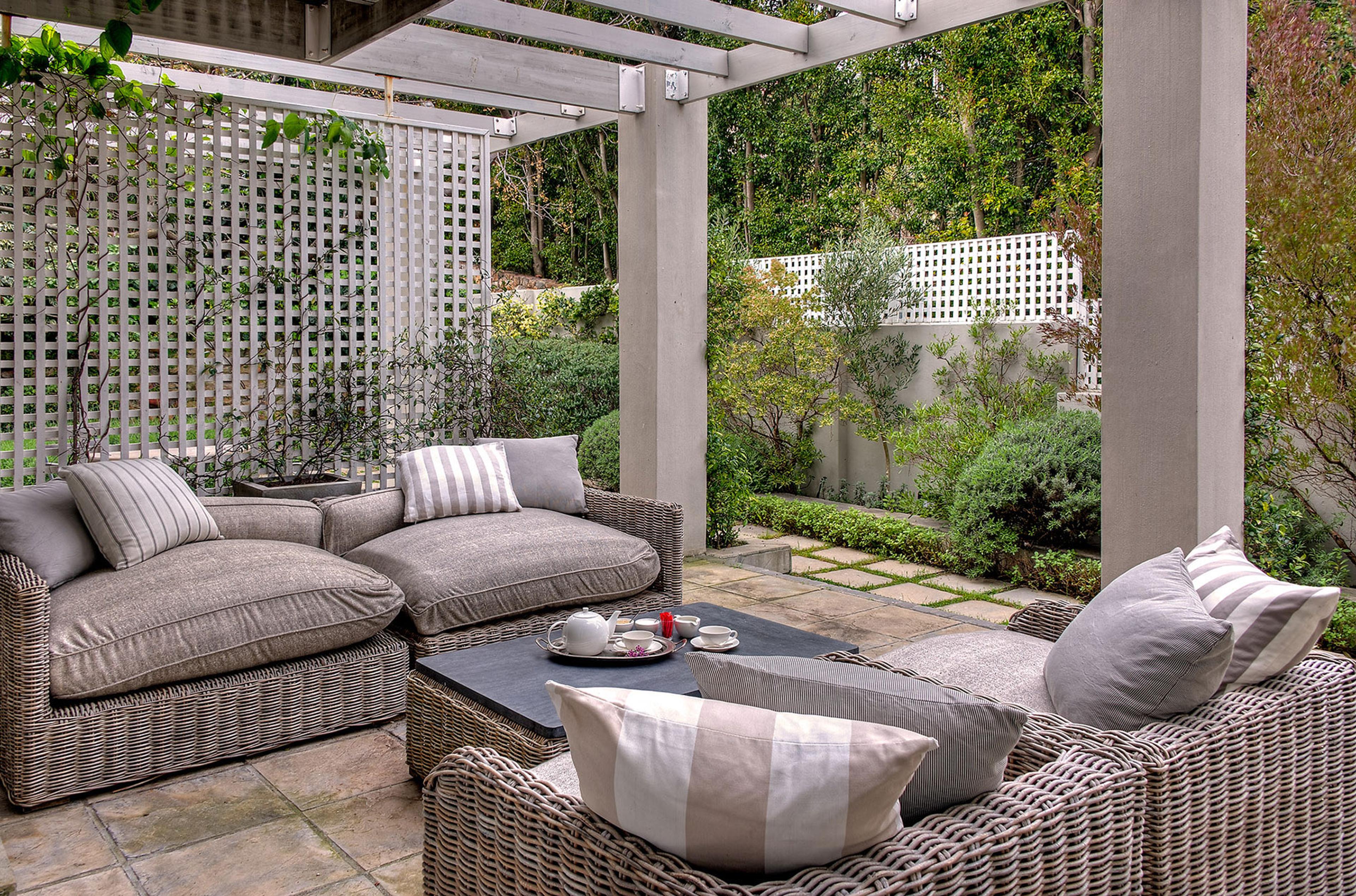 A covered pergola patio with grey wicker sofas, striped cushions and a low table with a tea tray, beside clipped shrubs and a white lattice wall.