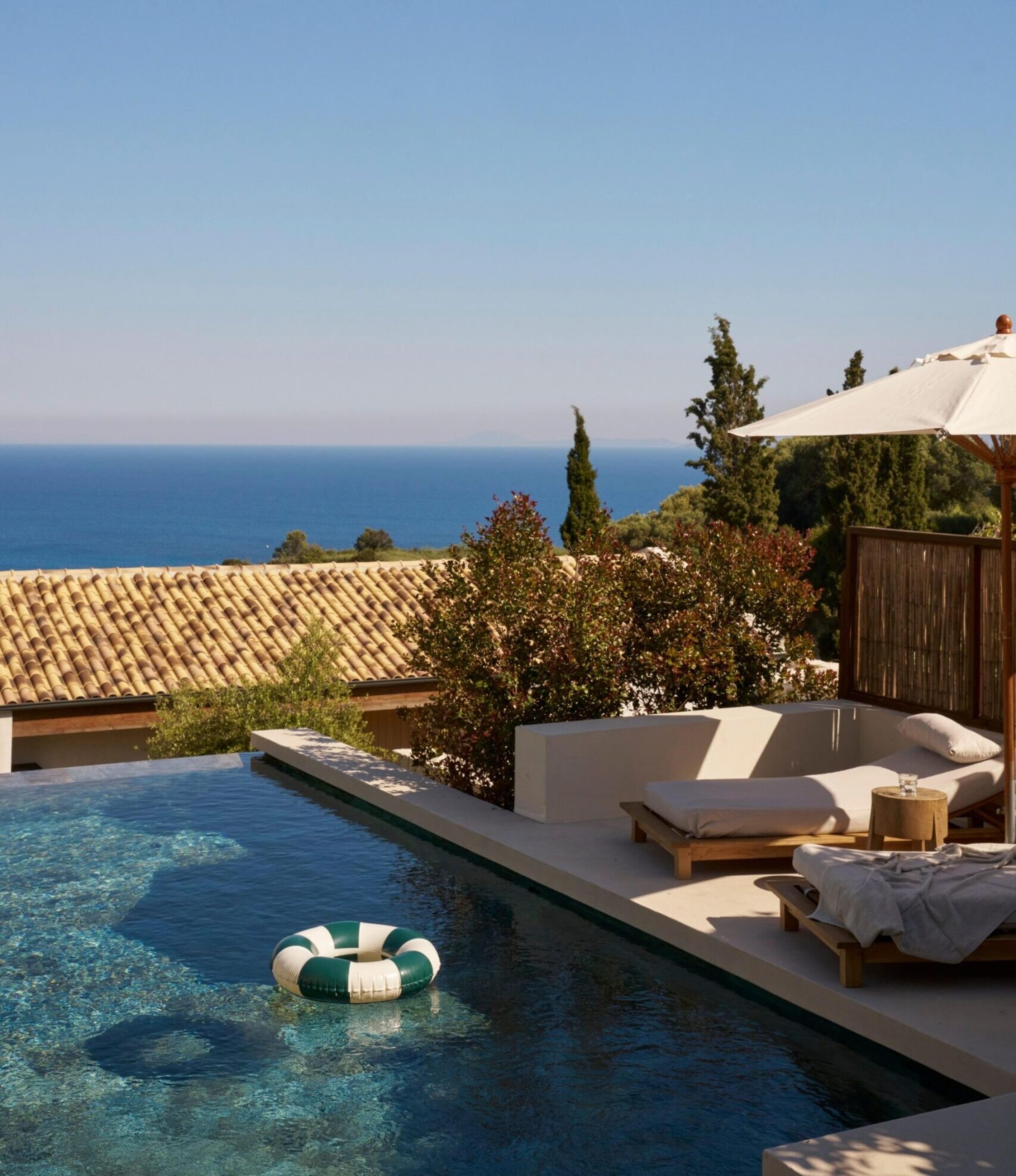 an infinity pool with a striped float on the water, sun loungers and a parasol beside it, the sea visible beyond a terracotta roofline
