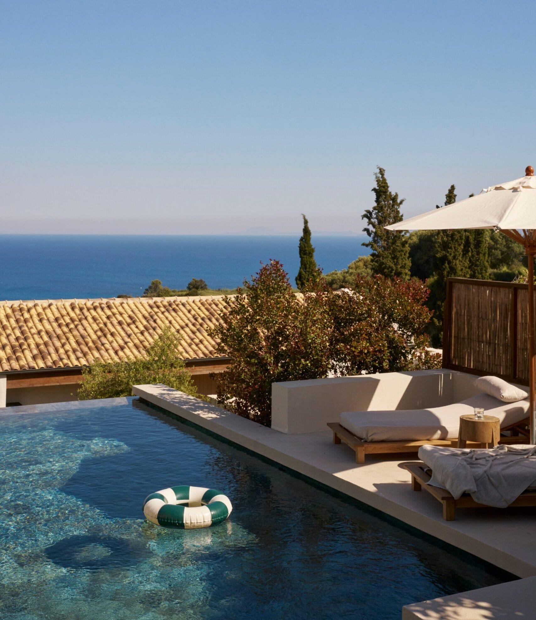 an infinity pool with a striped float on the water, sun loungers and a parasol beside it, the sea visible beyond a terracotta roofline