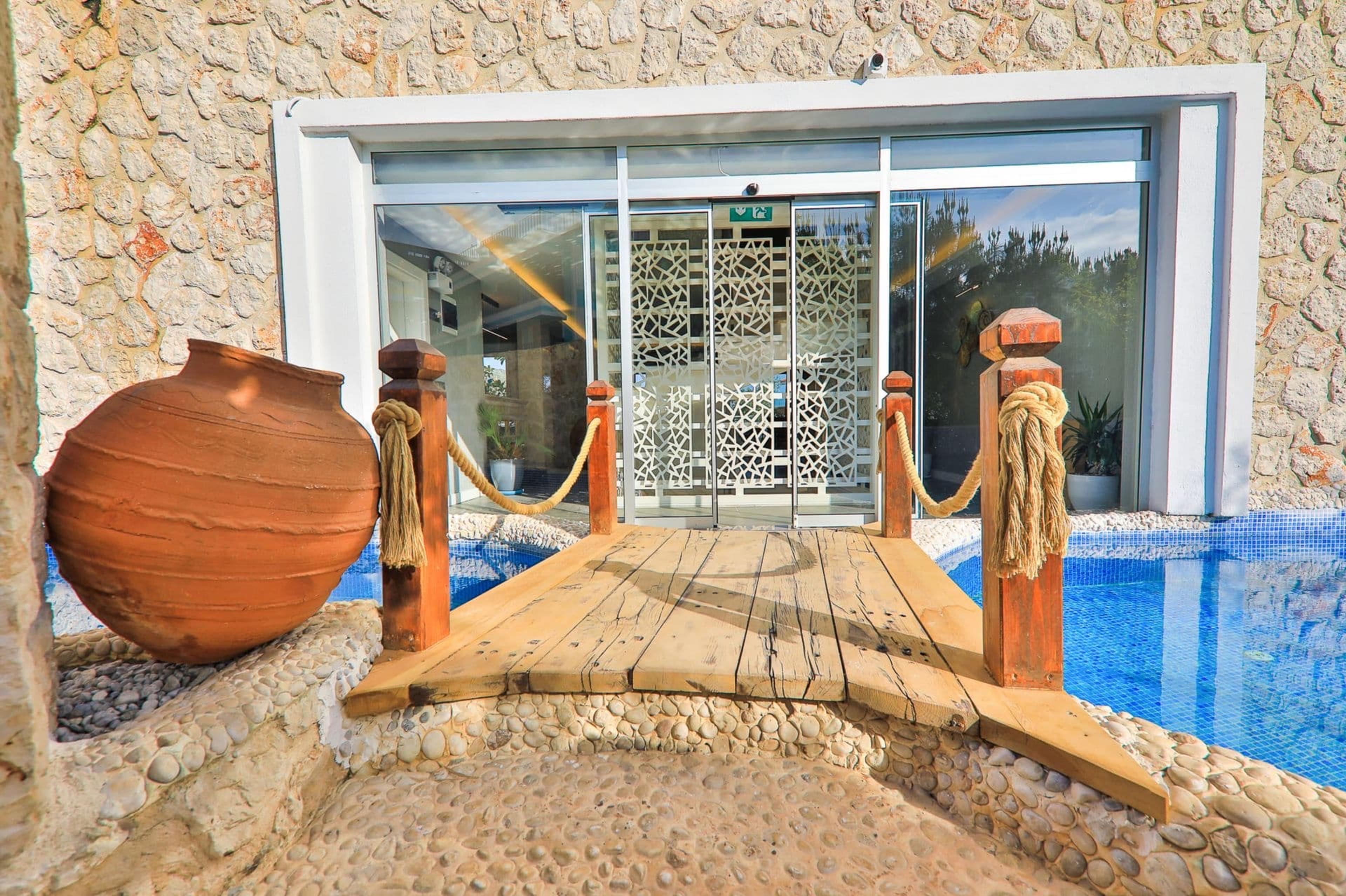 A wooden walkway crosses the edge of a swimming pool to glass doors, flanked by rope-wrapped posts and a large terracotta urn.