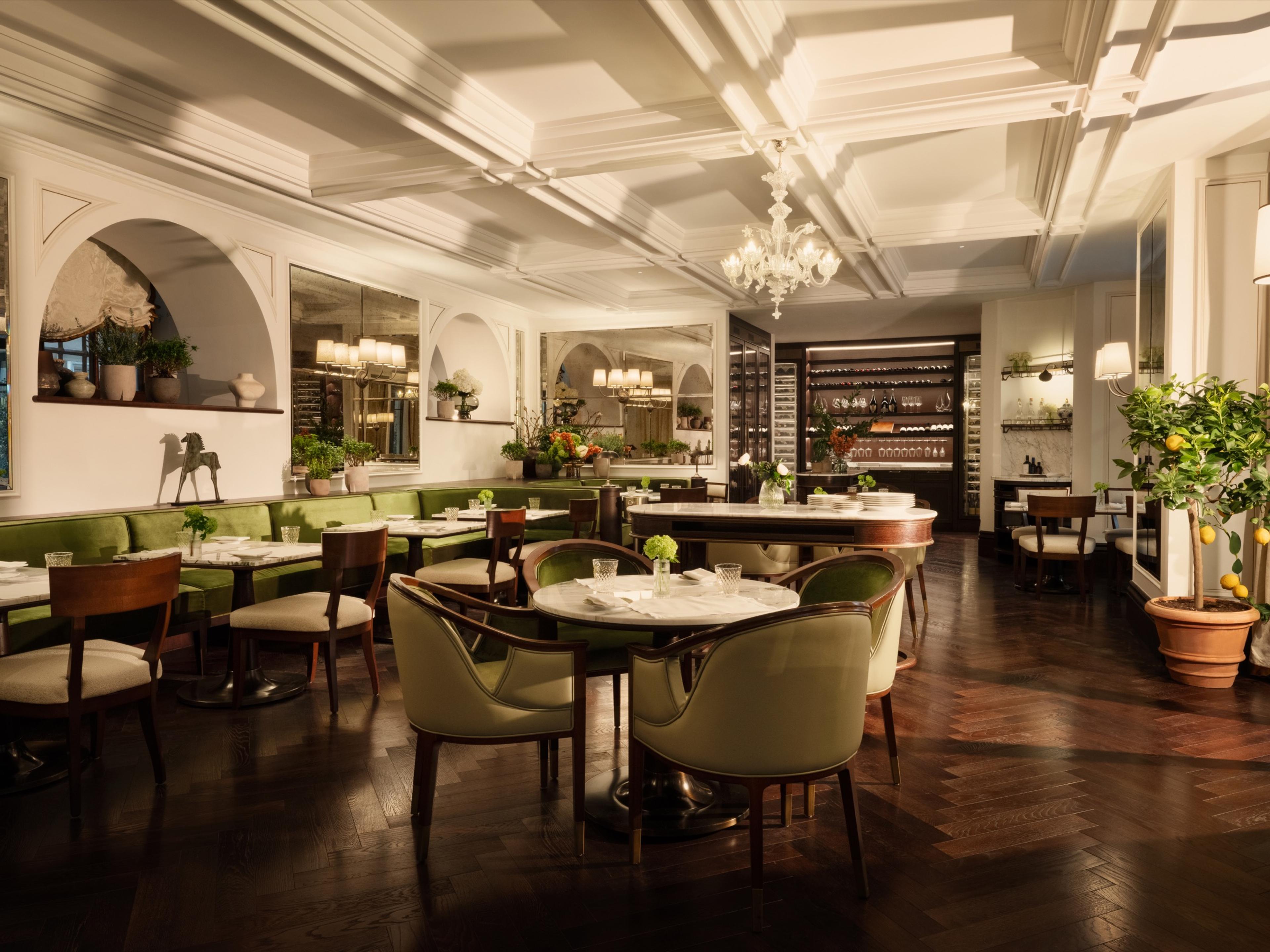 an empty restaurant dining room with green banquettes, round marble tables, a chandelier and a lemon tree in the corner