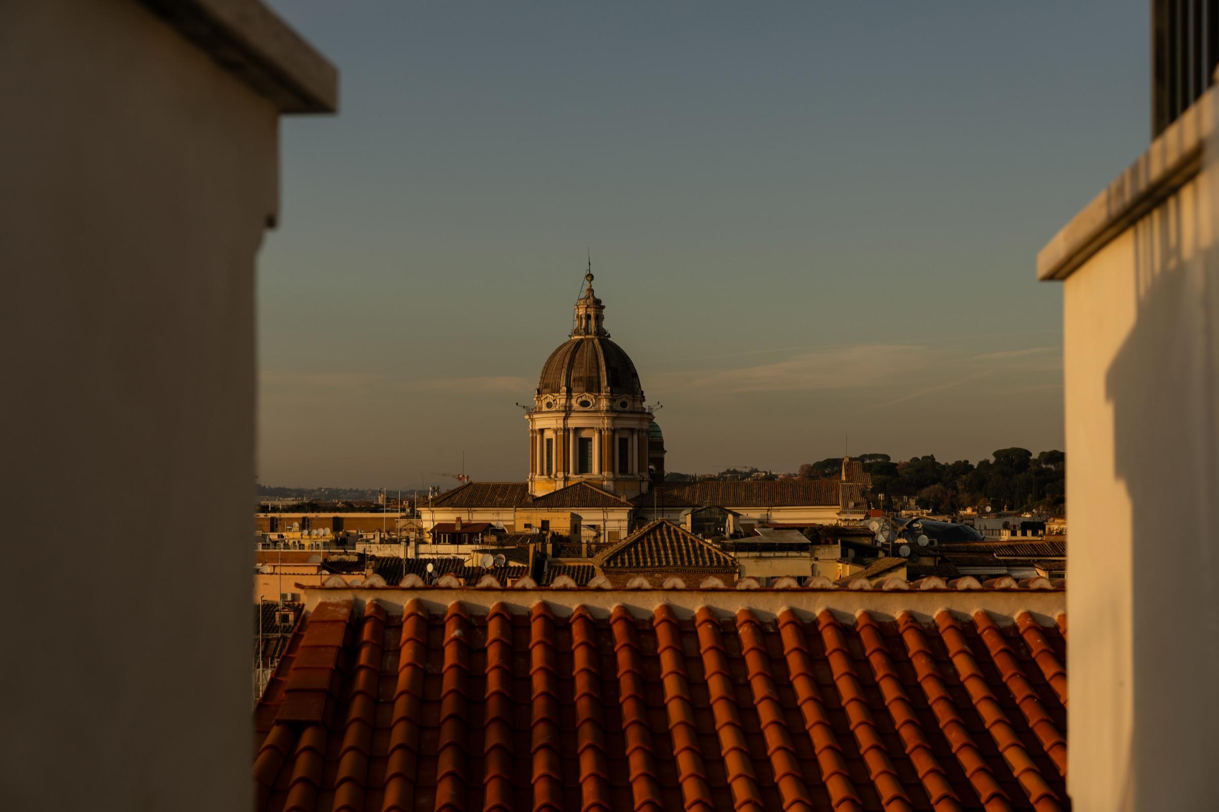 terracotta rooftops at dusk with a domed church rising above them, seen between two walls