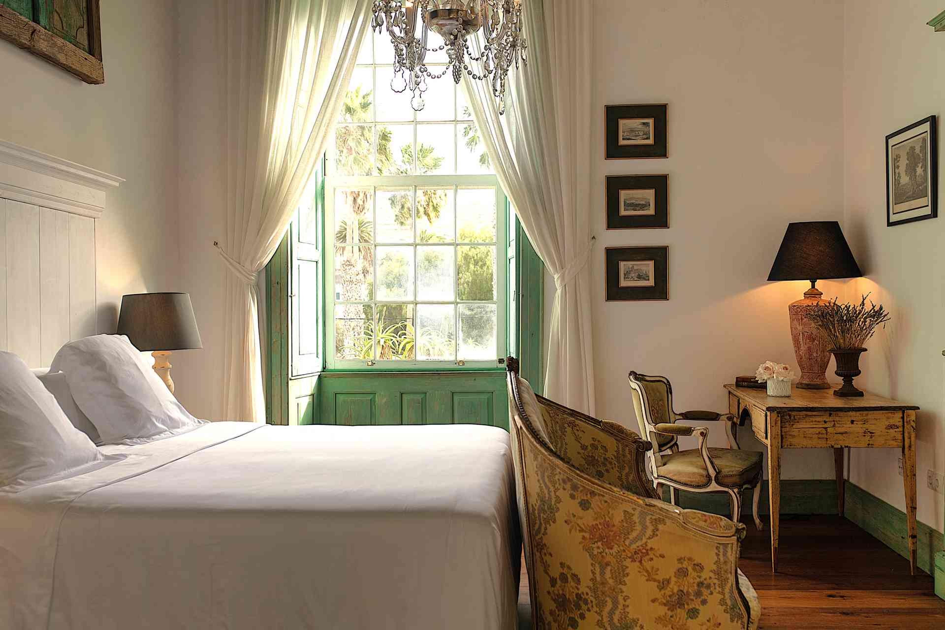 A bedroom with a white panelled headboard, a crystal chandelier, antique upholstered chairs and tall windows with green shutters open to the garden.