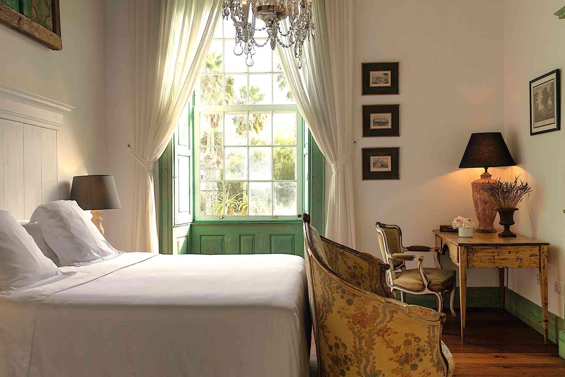 A bedroom with a white panelled headboard, a crystal chandelier, antique upholstered chairs and tall windows with green shutters open to the garden.