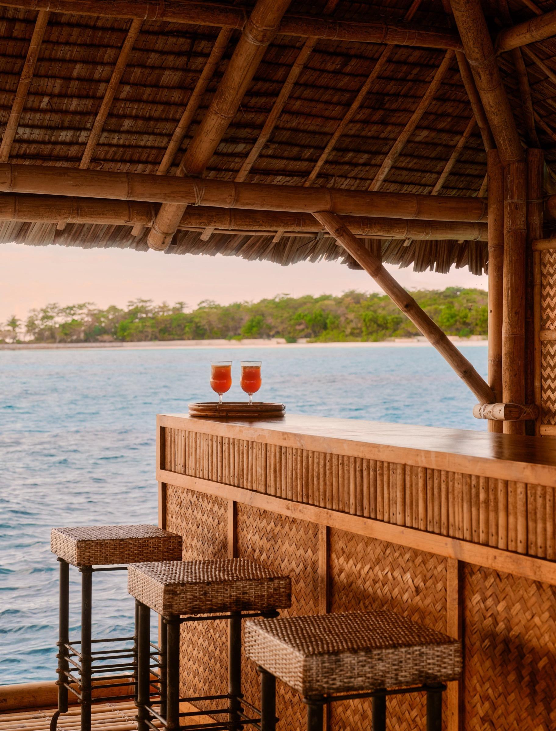 Two cocktails on a tray at a thatched roof bar built over the water, with trees on the shore beyond.