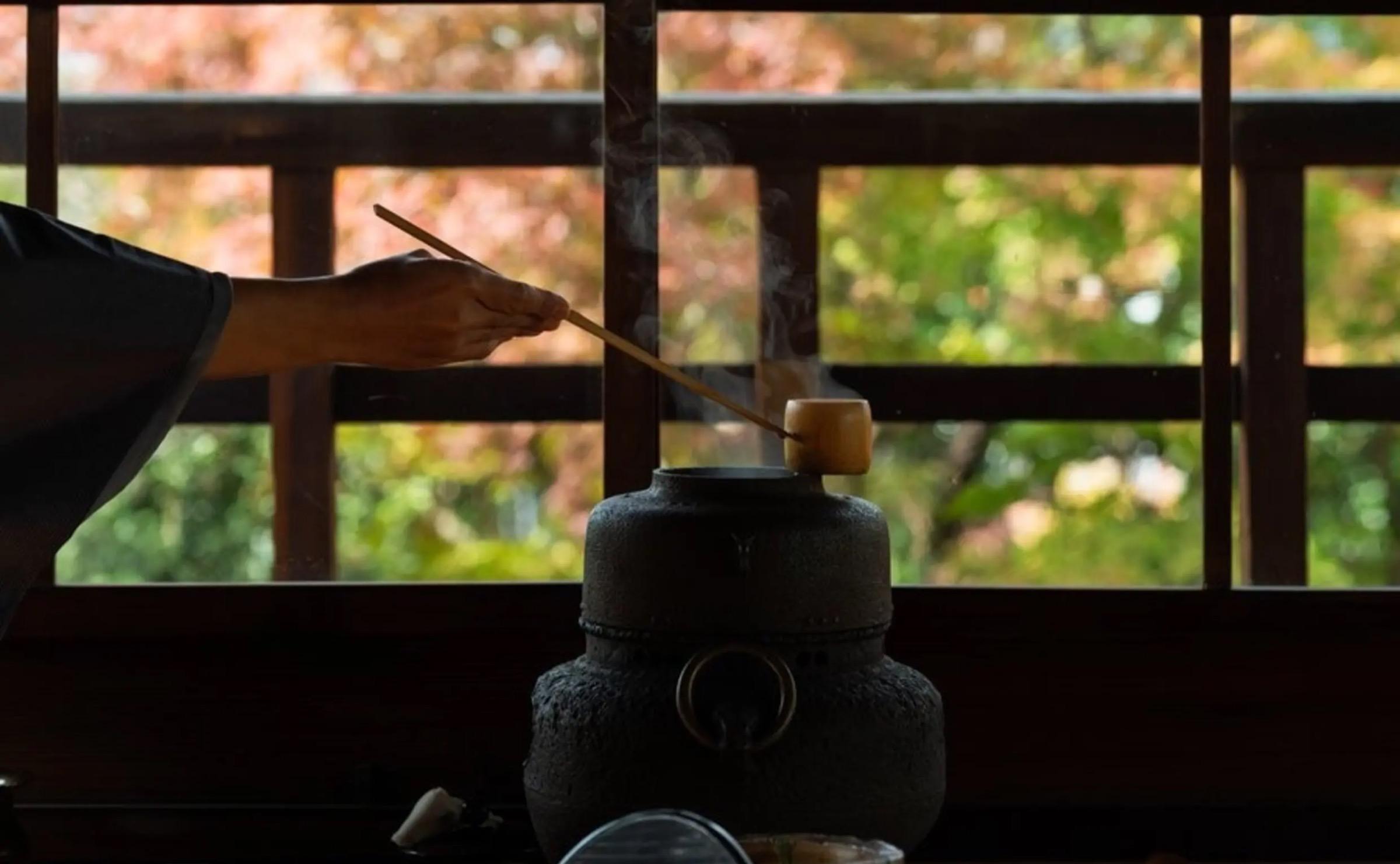 A hand holds a bamboo implement over a steaming black iron kettle, with autumn foliage blurred in the window behind.