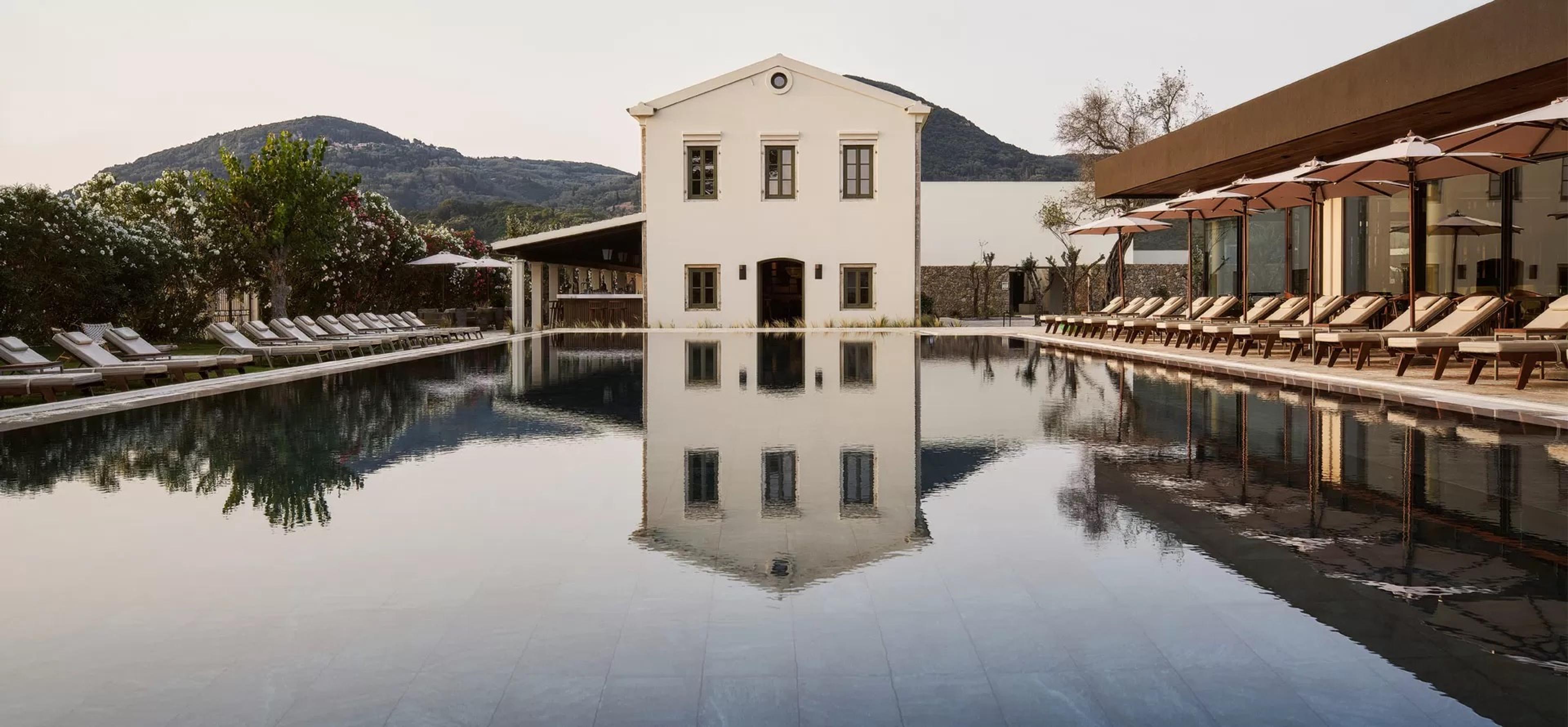 A rectangular pool reflecting a white two-storey building and rows of sun loungers, with hills behind at dusk.