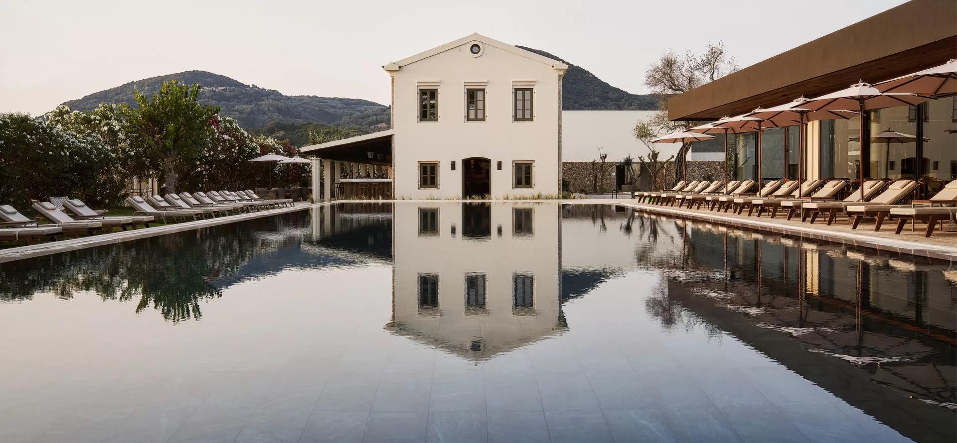A rectangular pool reflecting a white two-storey building and rows of sun loungers, with hills behind at dusk.