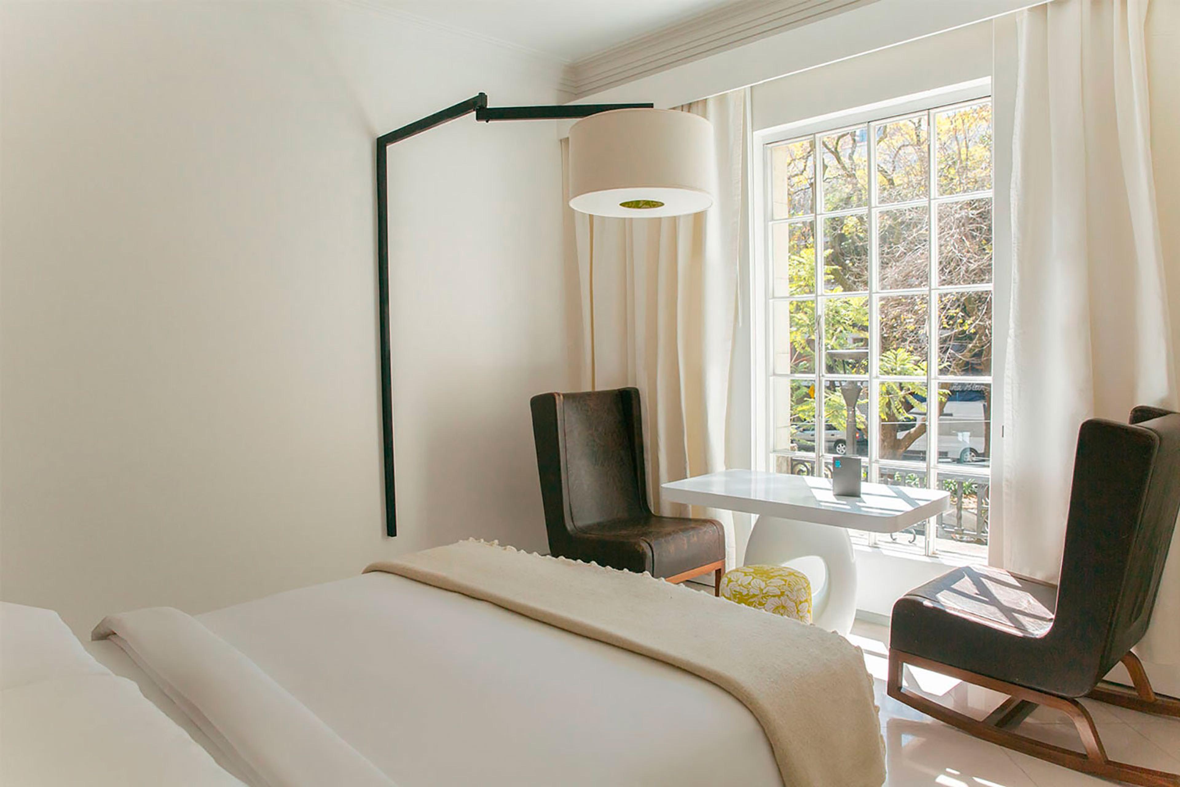 A bedroom with a cream bed, two brown leather chairs and a small table by a tall window onto a tree-lined street, with an angular floor lamp overhead.