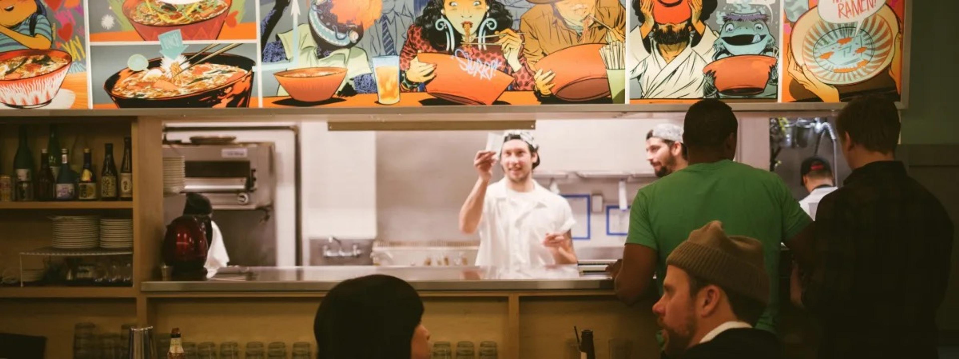 Cooks at an open kitchen counter under a bright painted mural, diners in the foreground