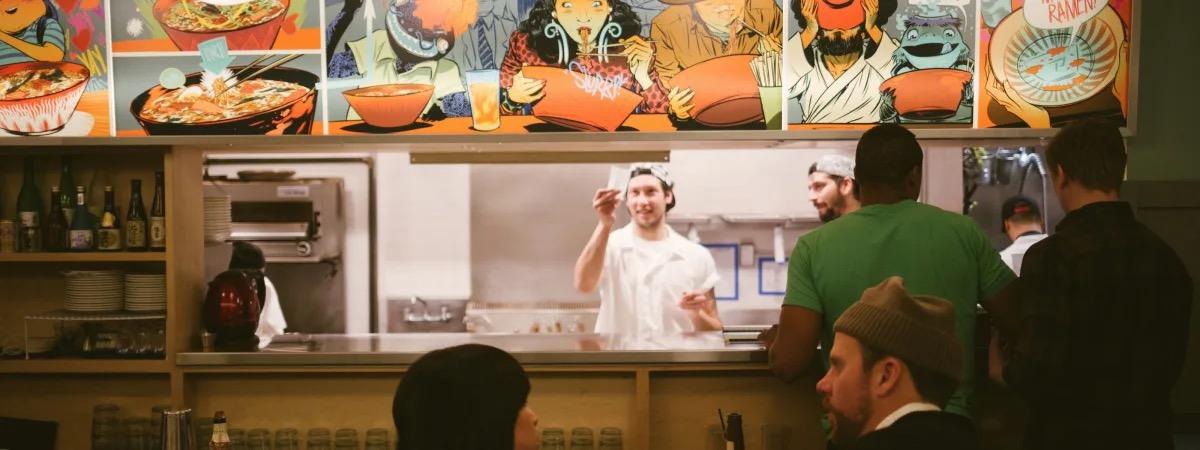 Cooks at an open kitchen counter under a bright painted mural, diners in the foreground