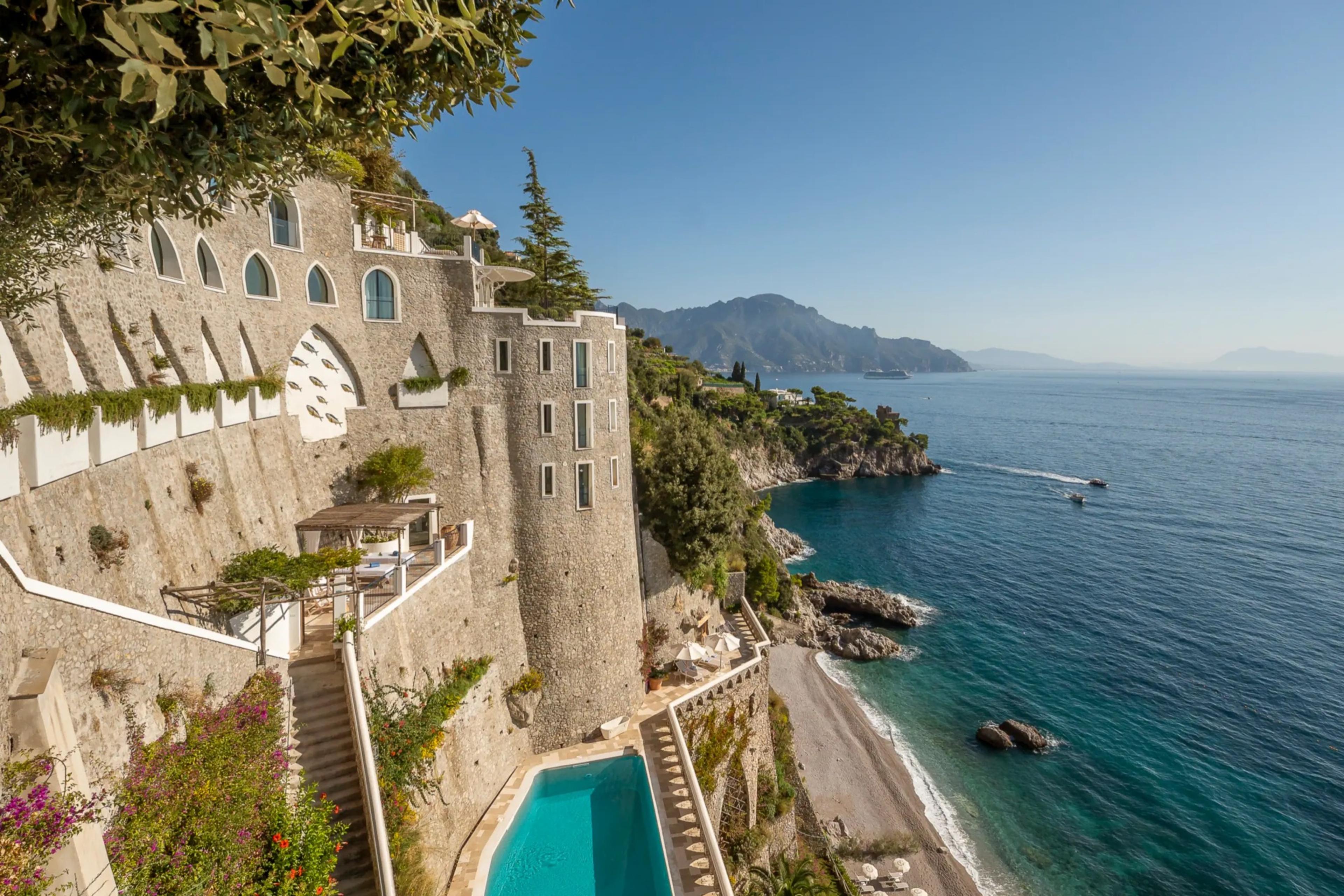 A multi-storey stone and white-plaster hotel built into a cliff face above the sea, with a swimming pool terrace at its base and pine branches framing the top of the frame.