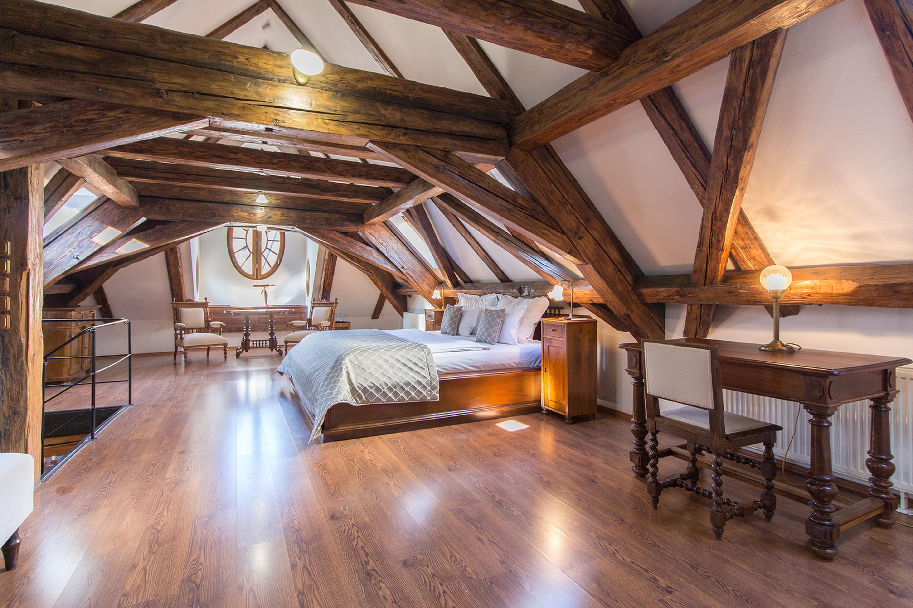 A bed and writing desk beneath a steeply pitched roof of exposed timber trusses.
