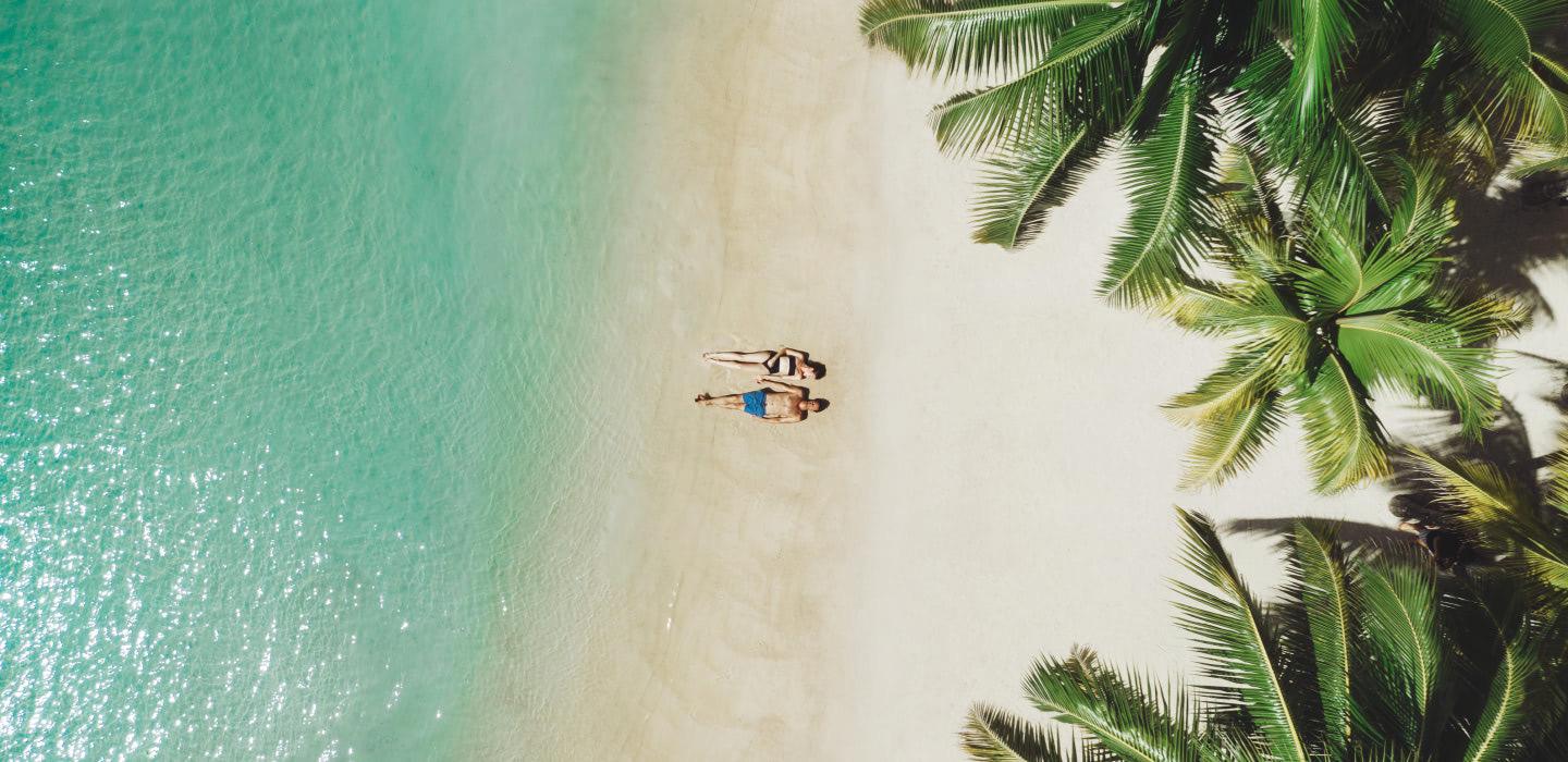 An aerial view of two people lying at the edge of a turquoise sea on white sand, with palm fronds framing one corner of the shot.