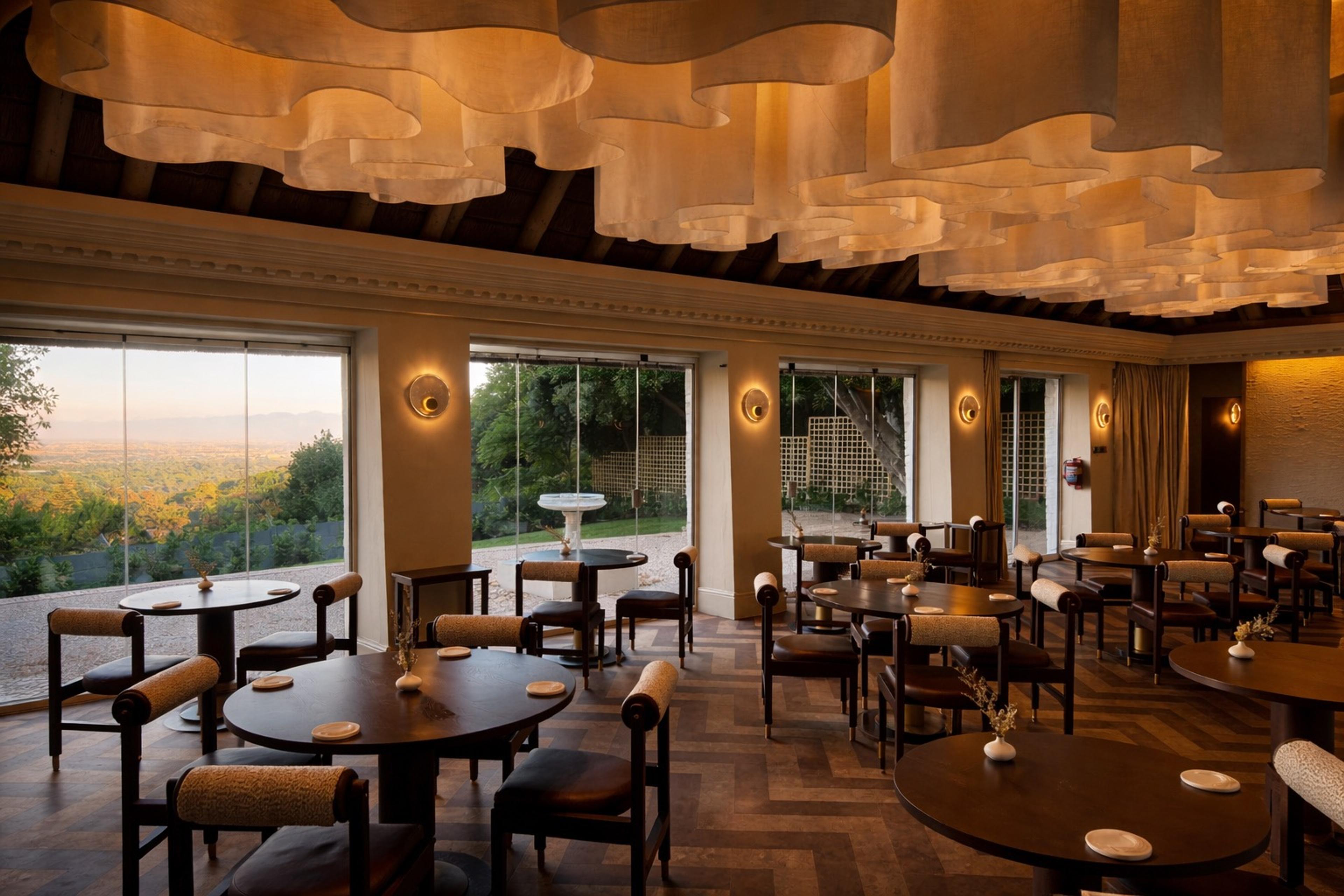 A dining room of wooden tables under pale hanging lampshades, with tall windows onto trees