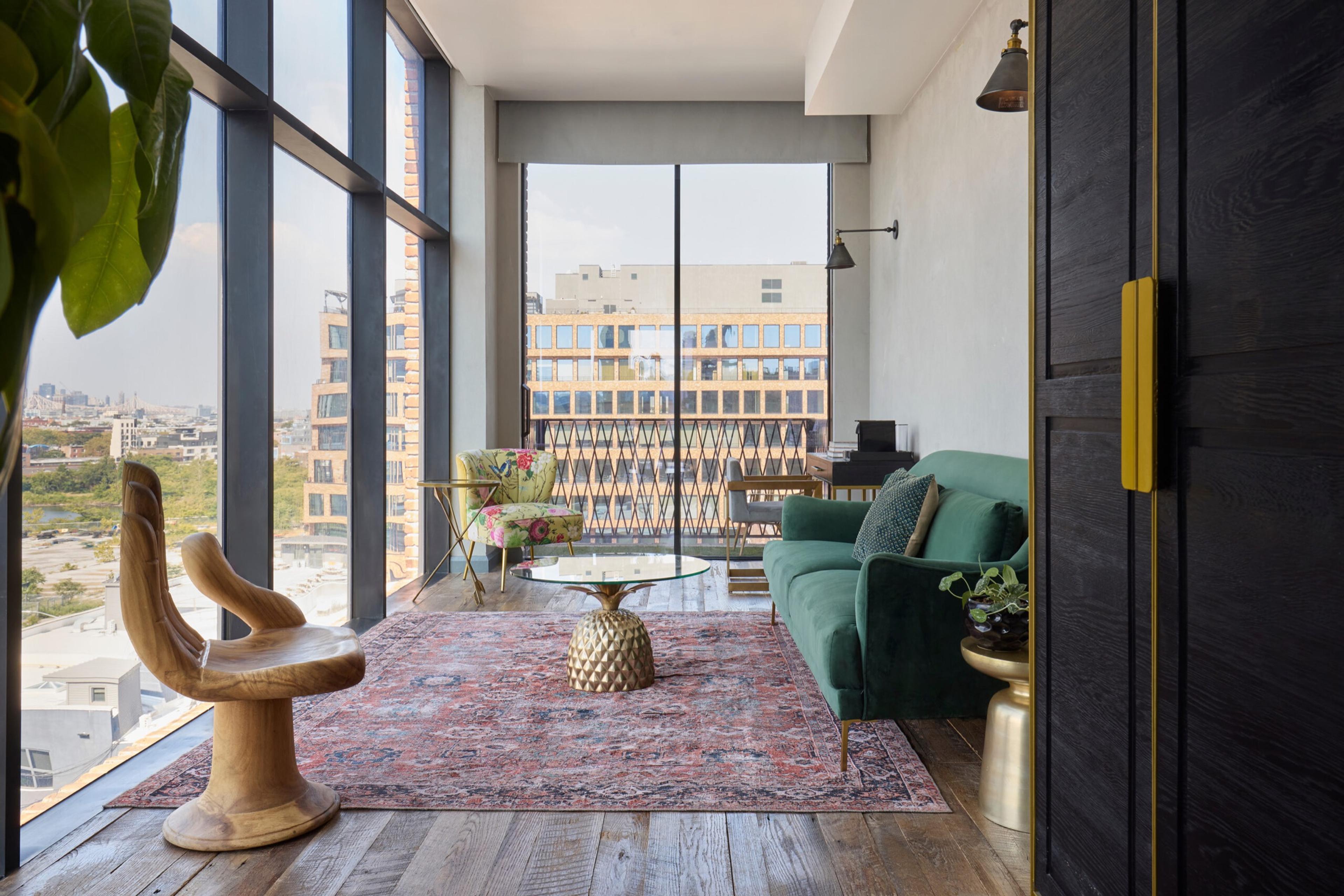 A suite living area with a sculptural wooden chair, a green sofa, a patterned rug, and floor-to-ceiling windows looking over nearby rooftops.