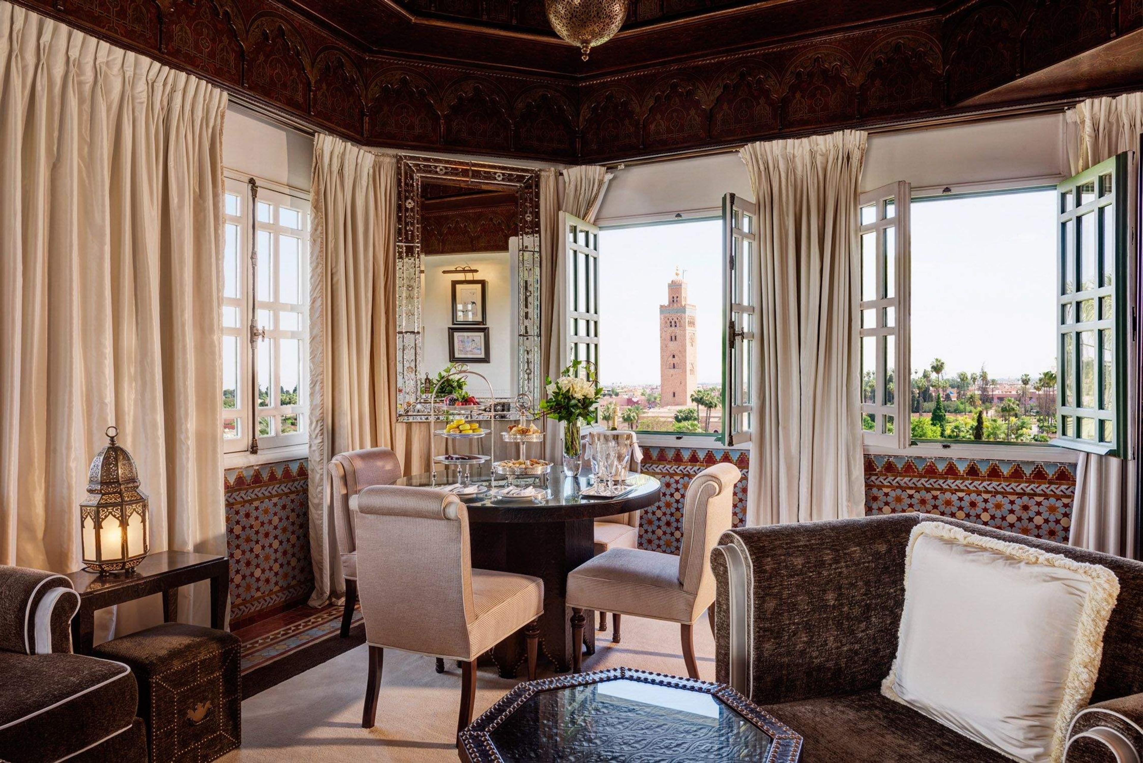 a sitting room with a carved wooden ceiling and lantern, armchairs around a table set with fruit and flowers, and open windows framing the Koutoubia minaret beyond the gardens