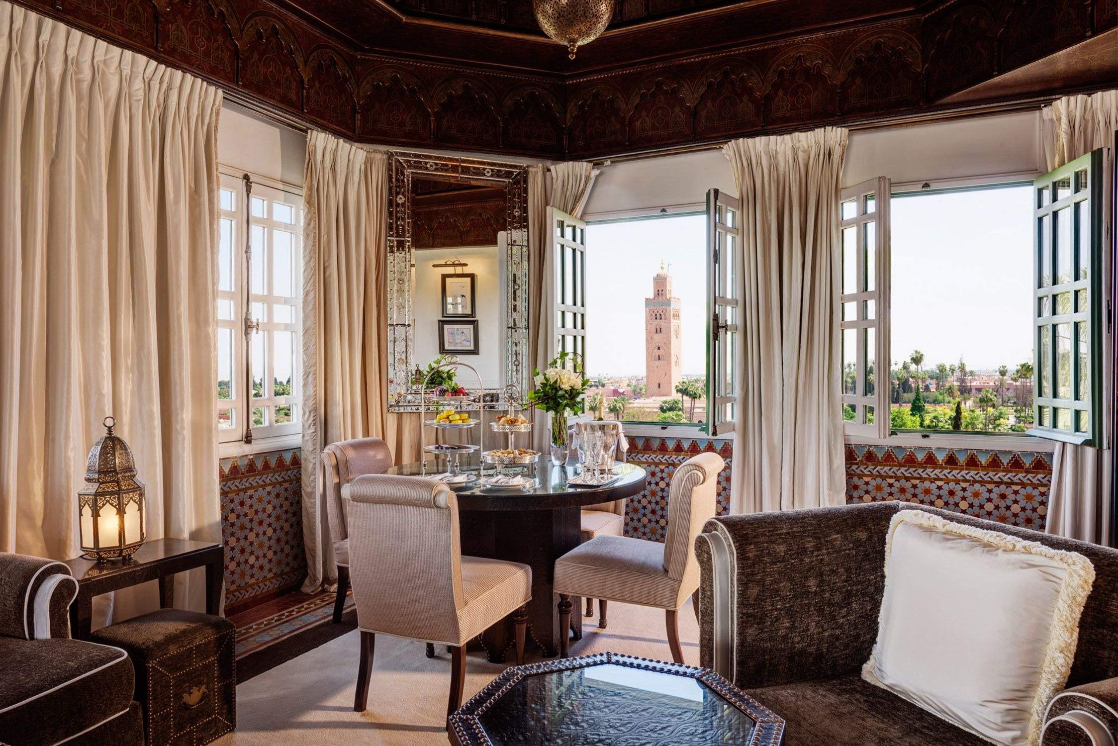 a sitting room with a carved wooden ceiling and lantern, armchairs around a table set with fruit and flowers, and open windows framing the Koutoubia minaret beyond the gardens