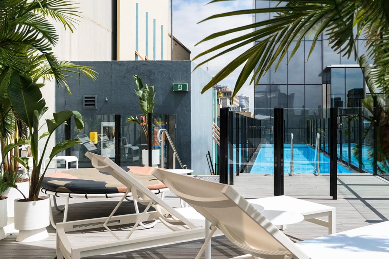 A rooftop terrace with cream sun loungers and potted palms beside a lap pool, enclosed by a glass balustrade with city buildings behind.