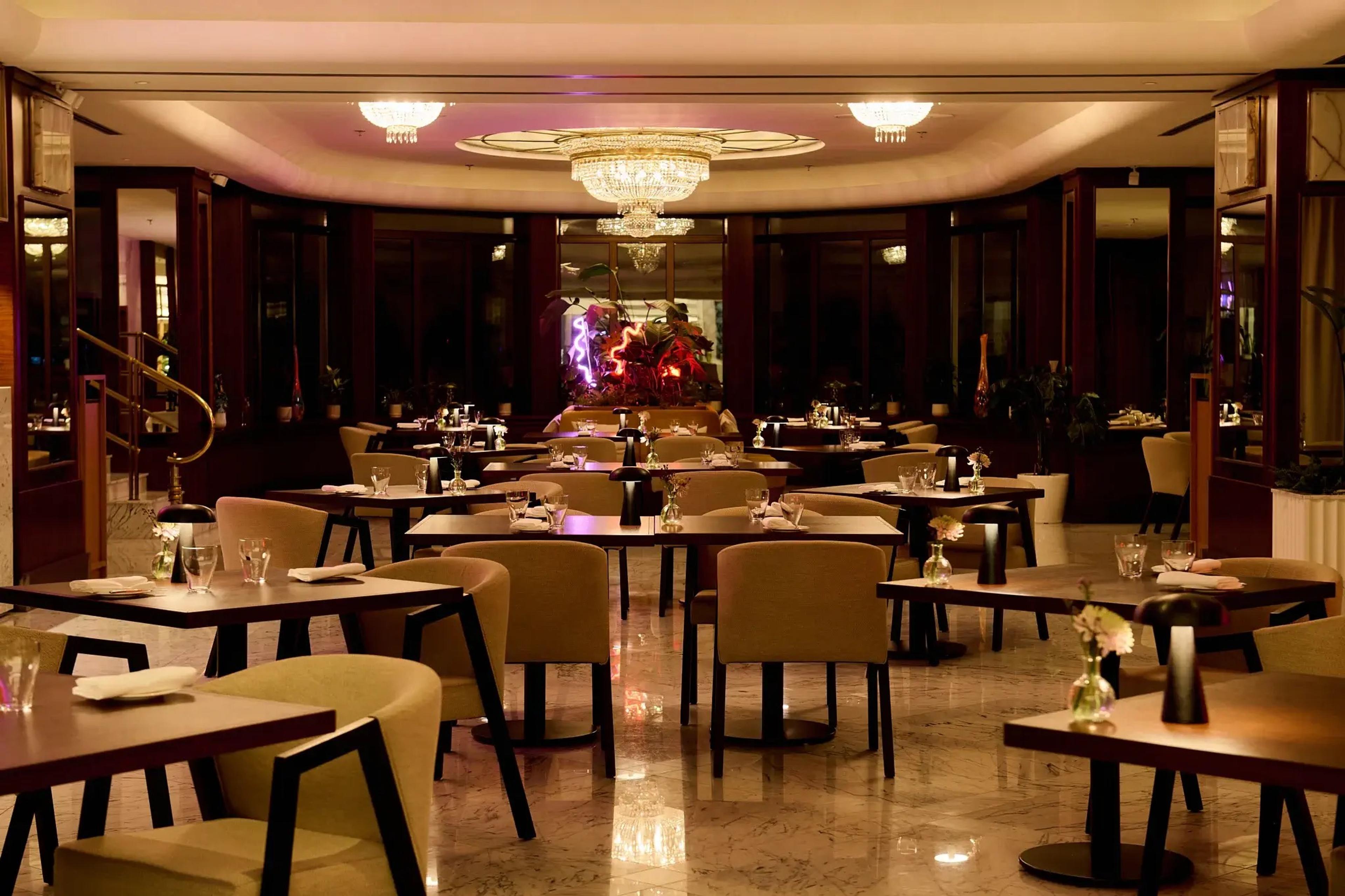 A dining room at night with tables laid for dinner, tan chairs, and a crystal chandelier over a central planting.
