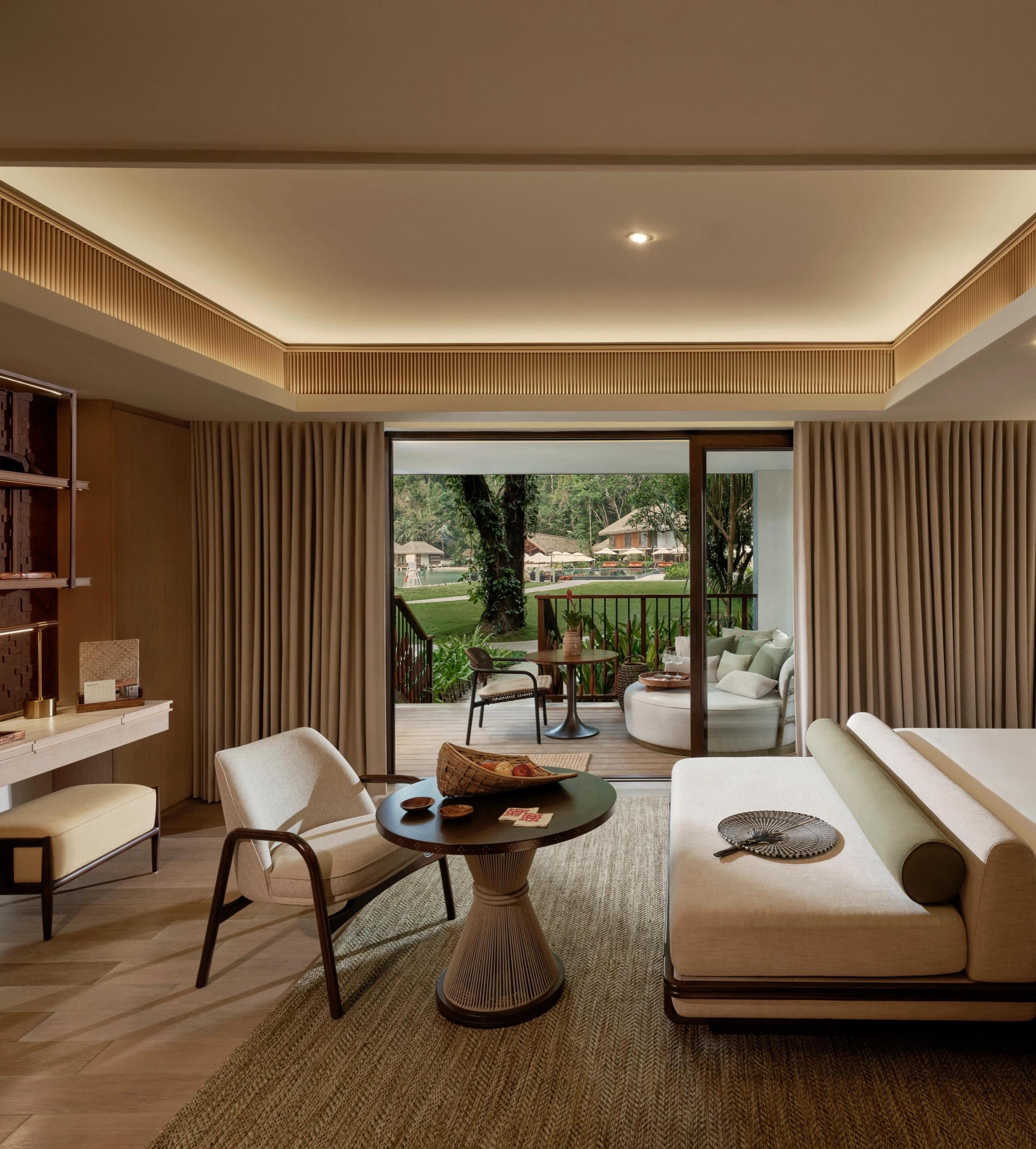 A bedroom with a low sofa and a desk, its glass doors opening onto a balcony among the trees.