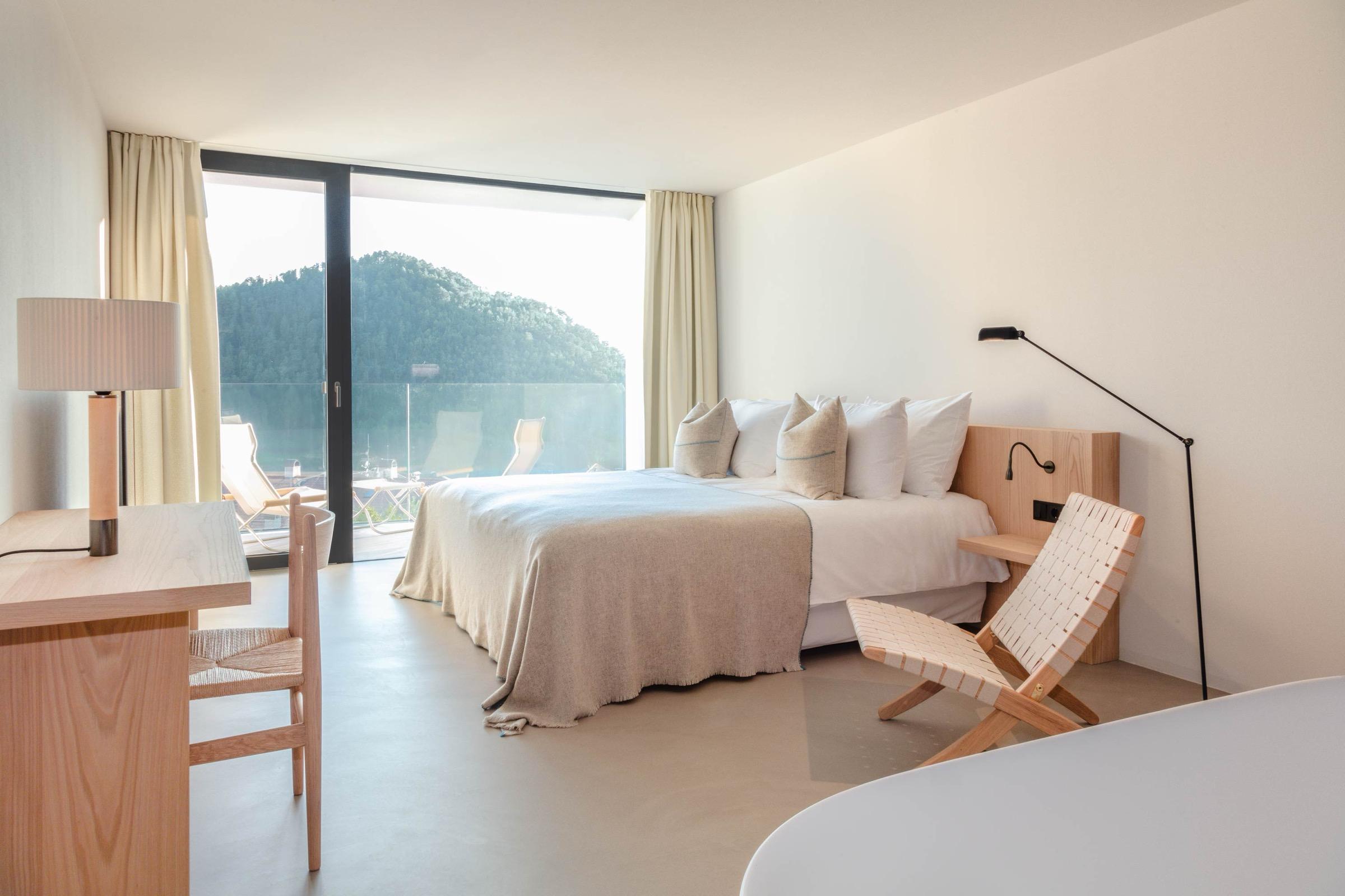 A hotel bed facing a floor-to-ceiling window onto a balcony with loungers, overlooking a green, tree-covered hillside.