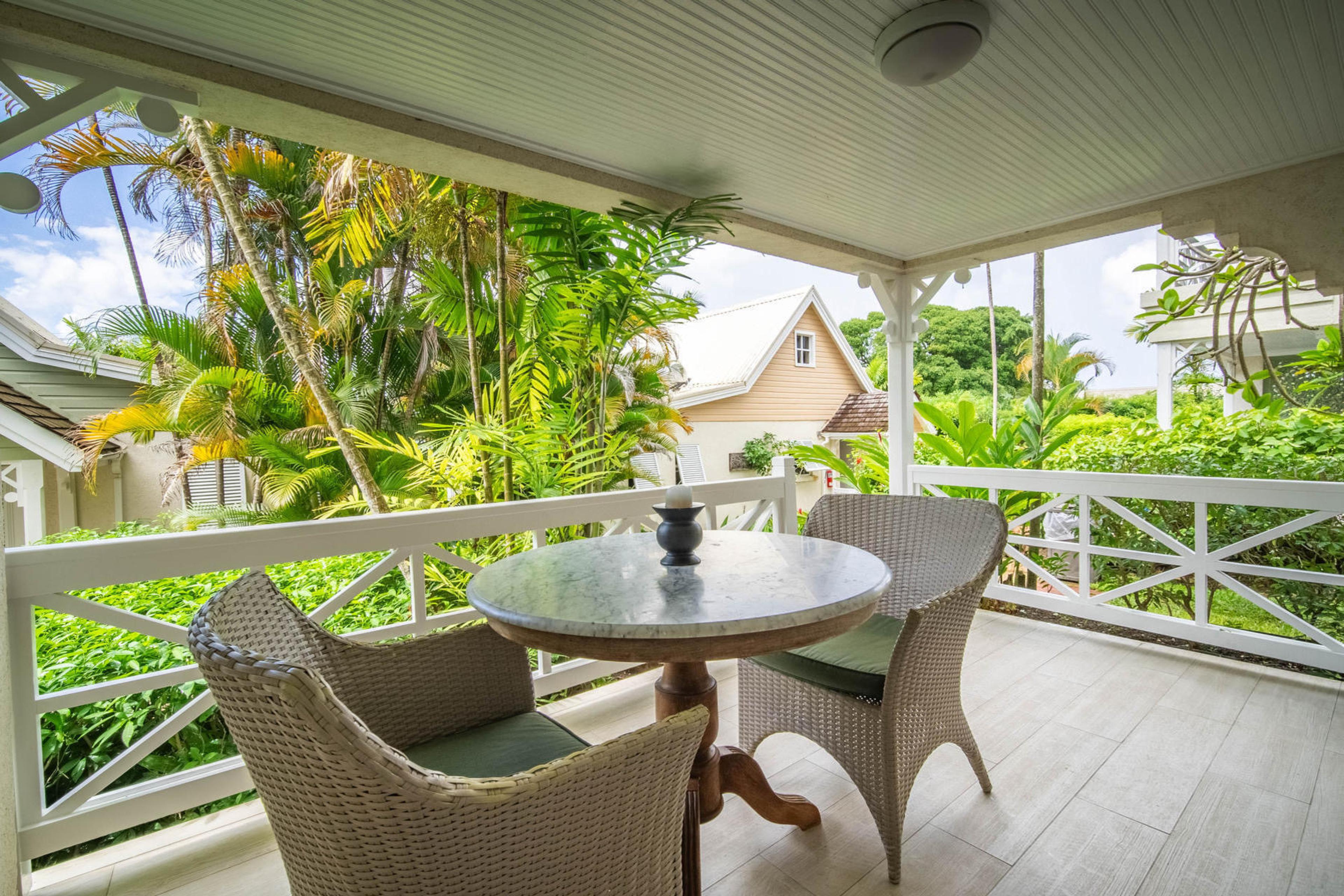 A covered balcony with a round stone table and two wicker chairs, overlooking palm trees and a pink cottage roof.