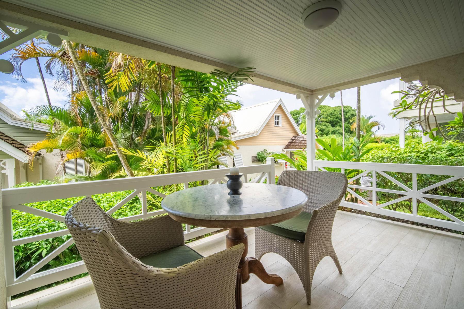 A covered balcony with a round stone table and two wicker chairs, overlooking palm trees and a pink cottage roof.