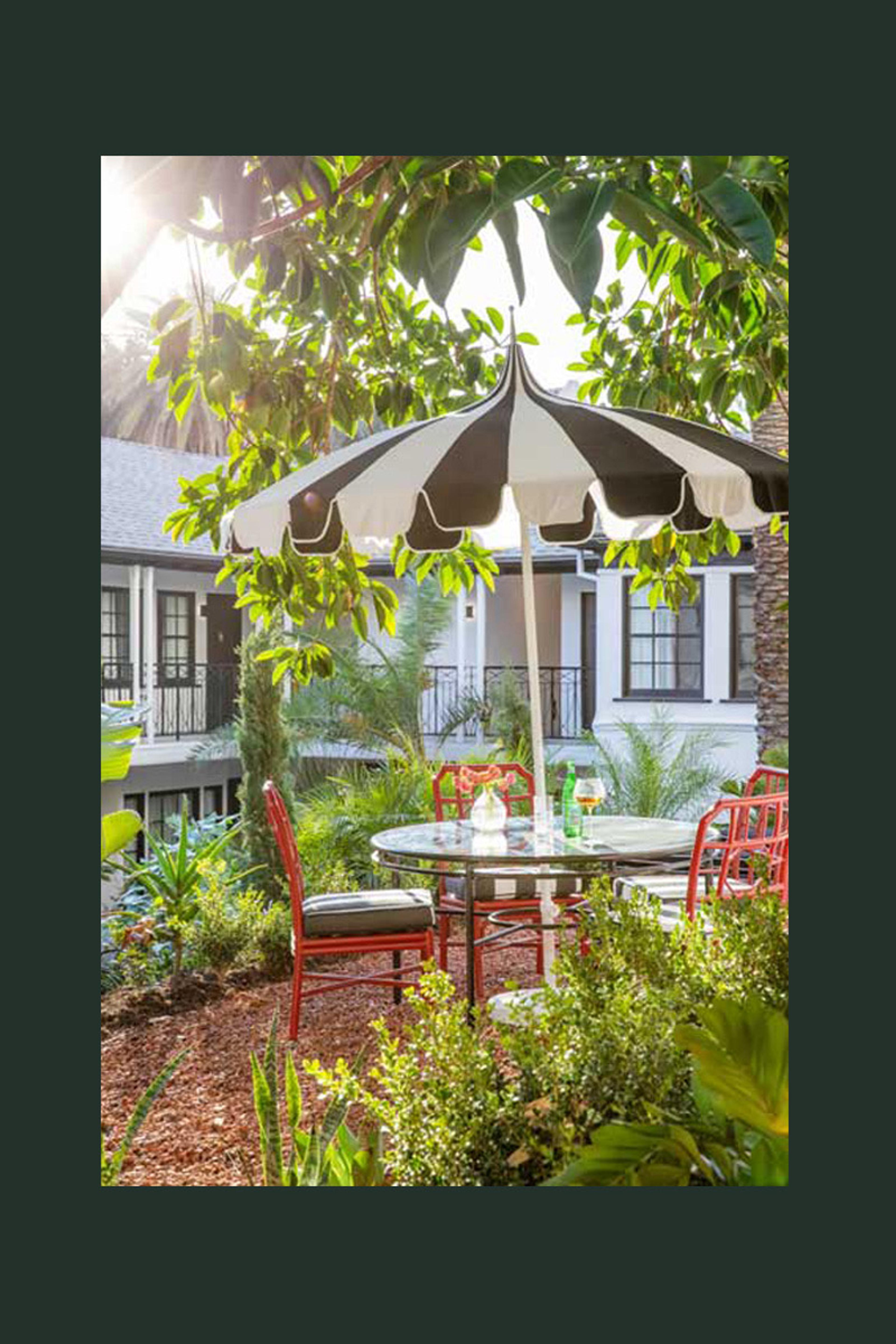 A round table set for two under a black and white striped umbrella in a garden, with red metal chairs and a white building with balconies behind.