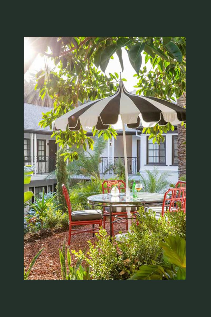 A round table set for two under a black and white striped umbrella in a garden, with red metal chairs and a white building with balconies behind.