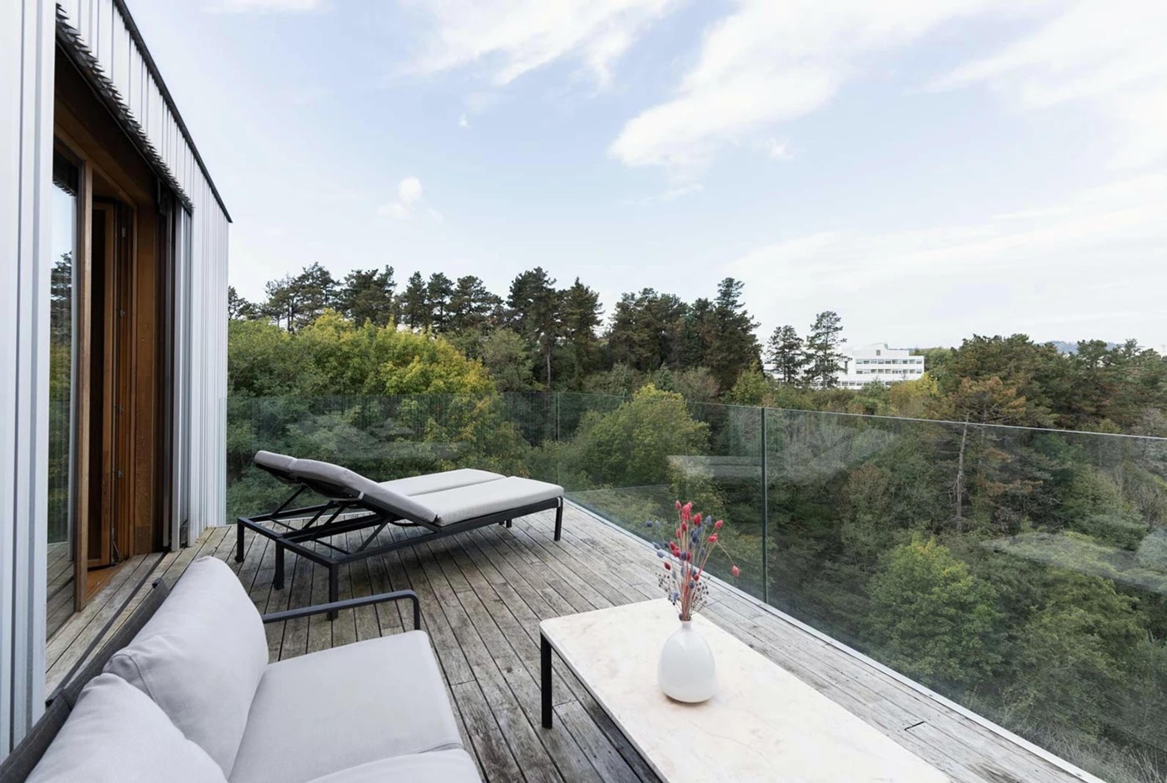A wooden rooftop terrace with a sunlounger and a sofa around a low table, enclosed by a glass balustrade, overlooking trees and a building under a partly cloudy sky.