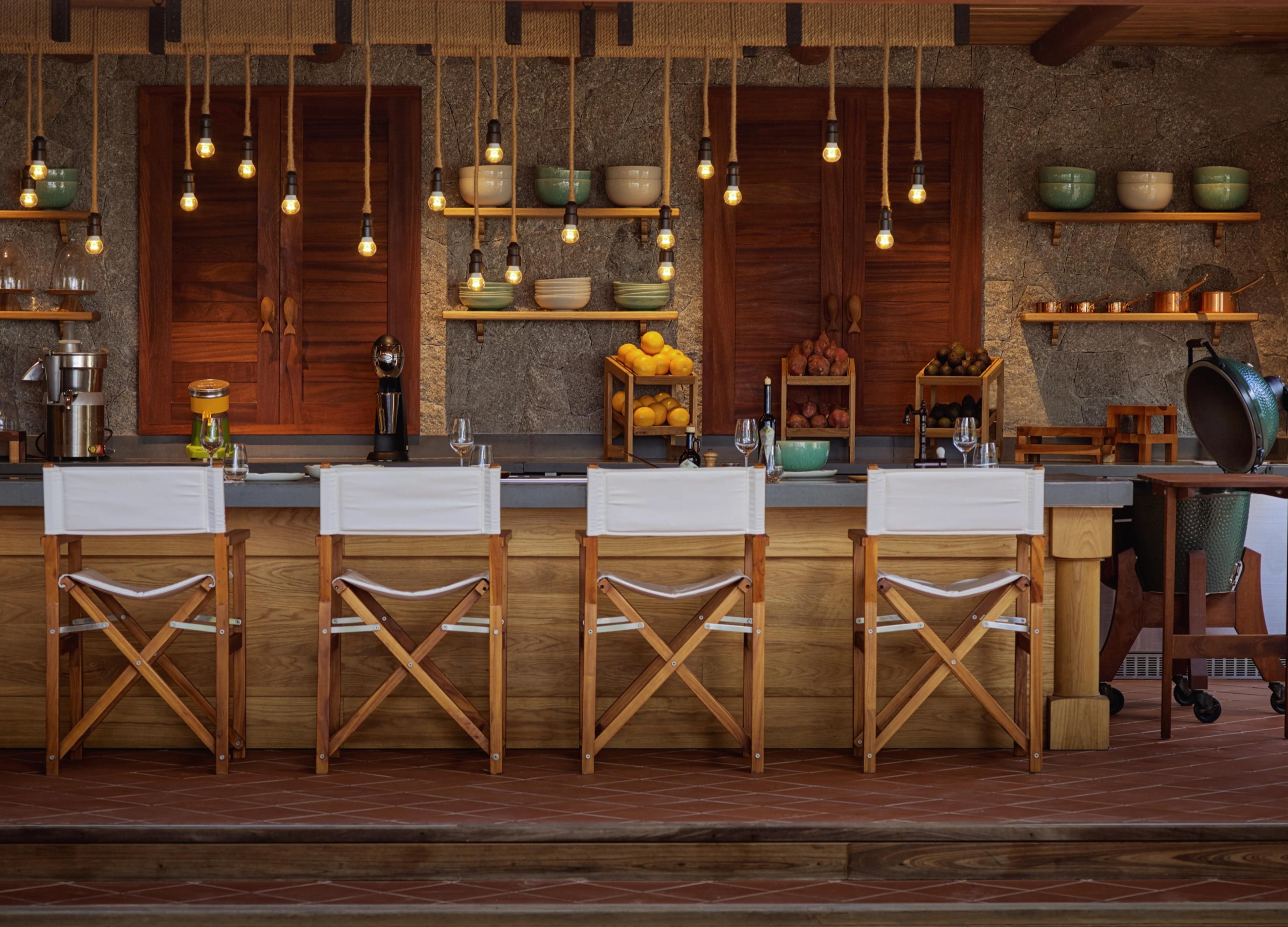 A stone kitchen counter with four director's chairs, hanging pendant lights, crates of oranges and bowls on open shelves.