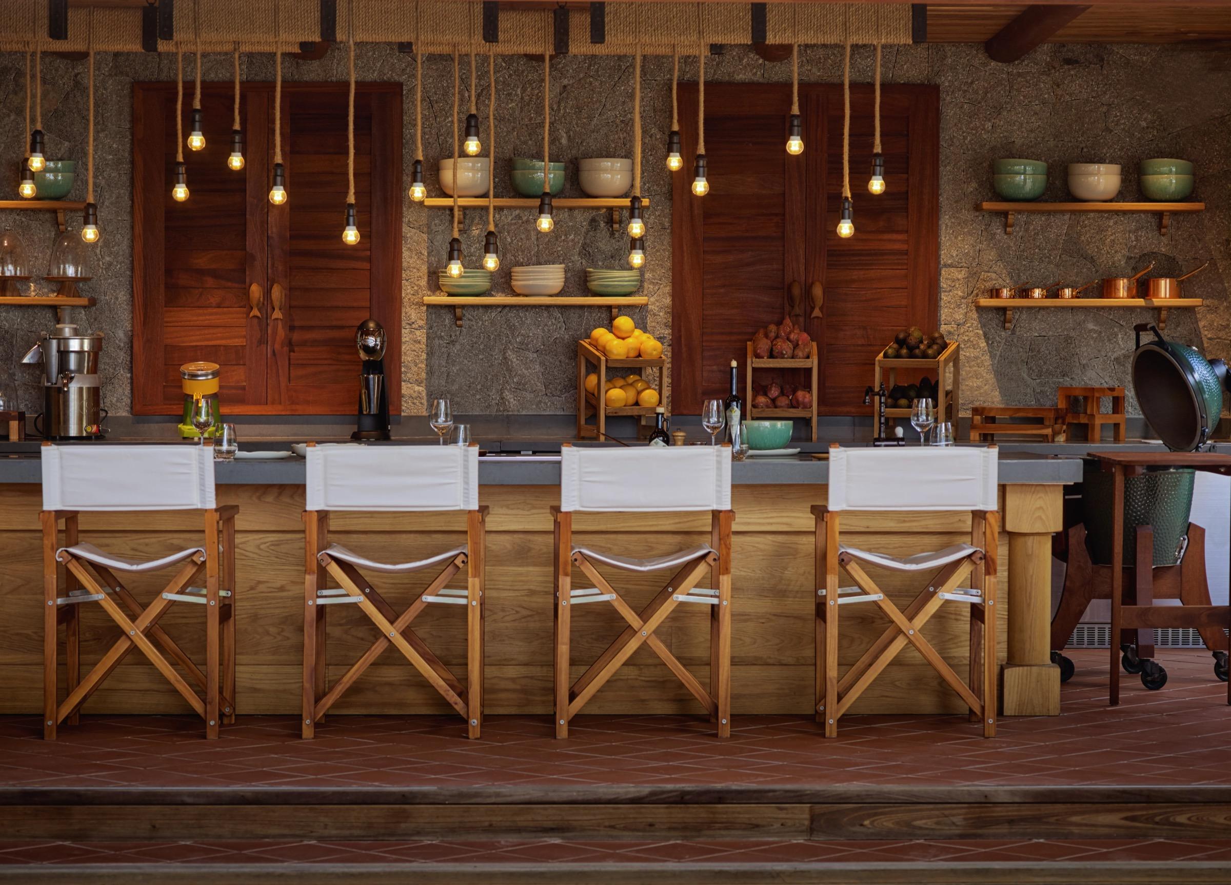 A stone kitchen counter with four director's chairs, hanging pendant lights, crates of oranges and bowls on open shelves.