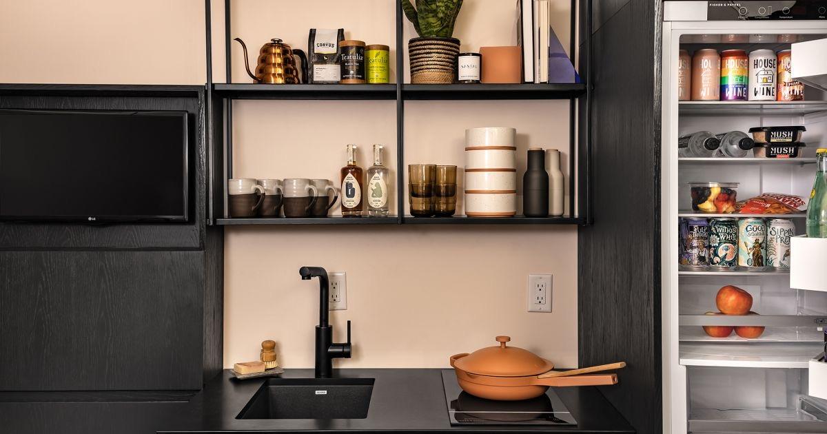 A hotel kitchenette shelf stocked with mugs, bottles and glassware beside a sink and induction hob, with a mini fridge stocked with canned drinks and fruit.