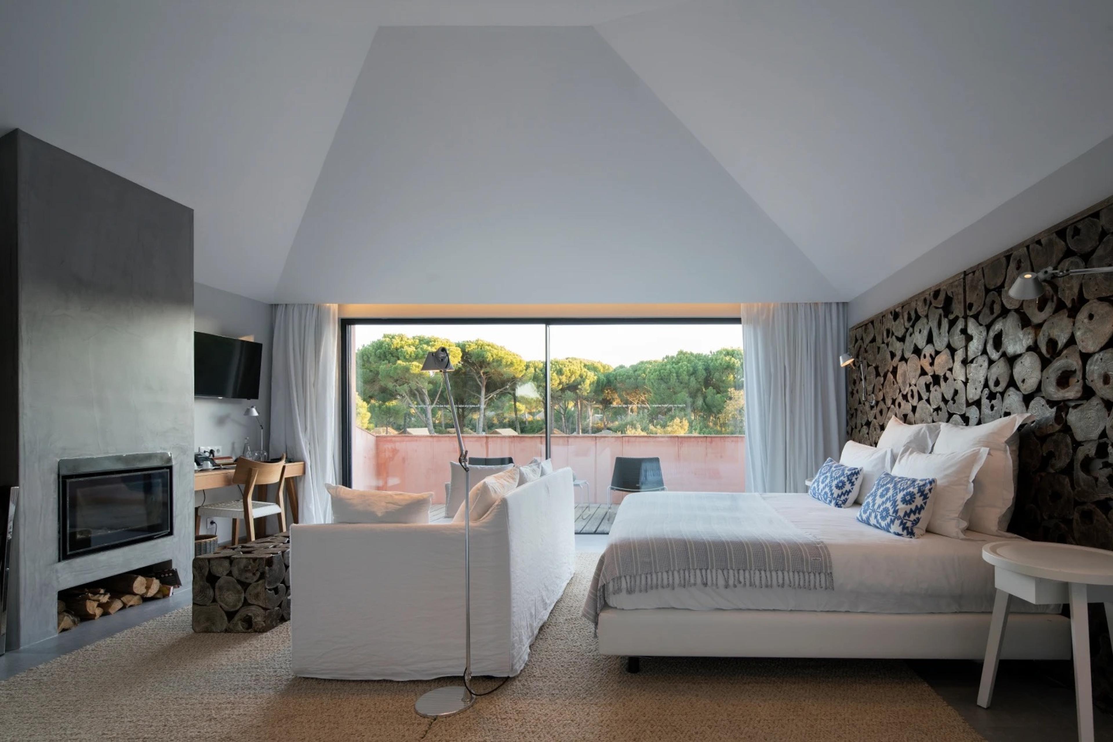 A bedroom with a vaulted ceiling, a reclaimed-wood headboard wall, a grey fireplace and a white sofa, with glass doors open to a terrace among pine trees.