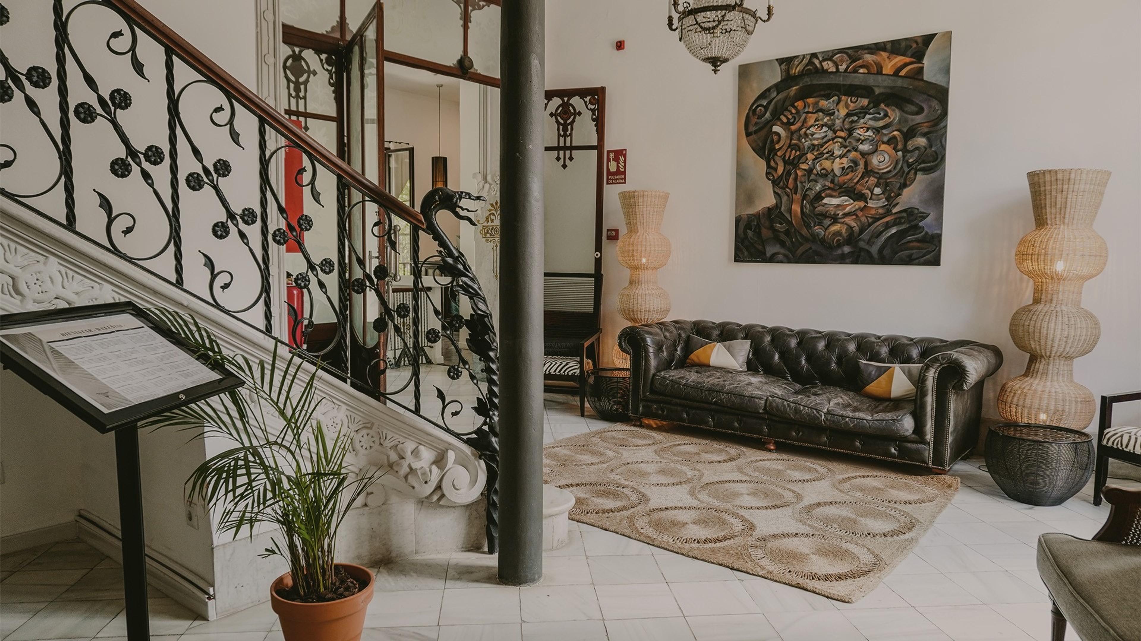 A hotel sitting room with a black leather Chesterfield sofa, a large painted portrait, woven floor lamps, an iron staircase railing, and a patterned rug.
