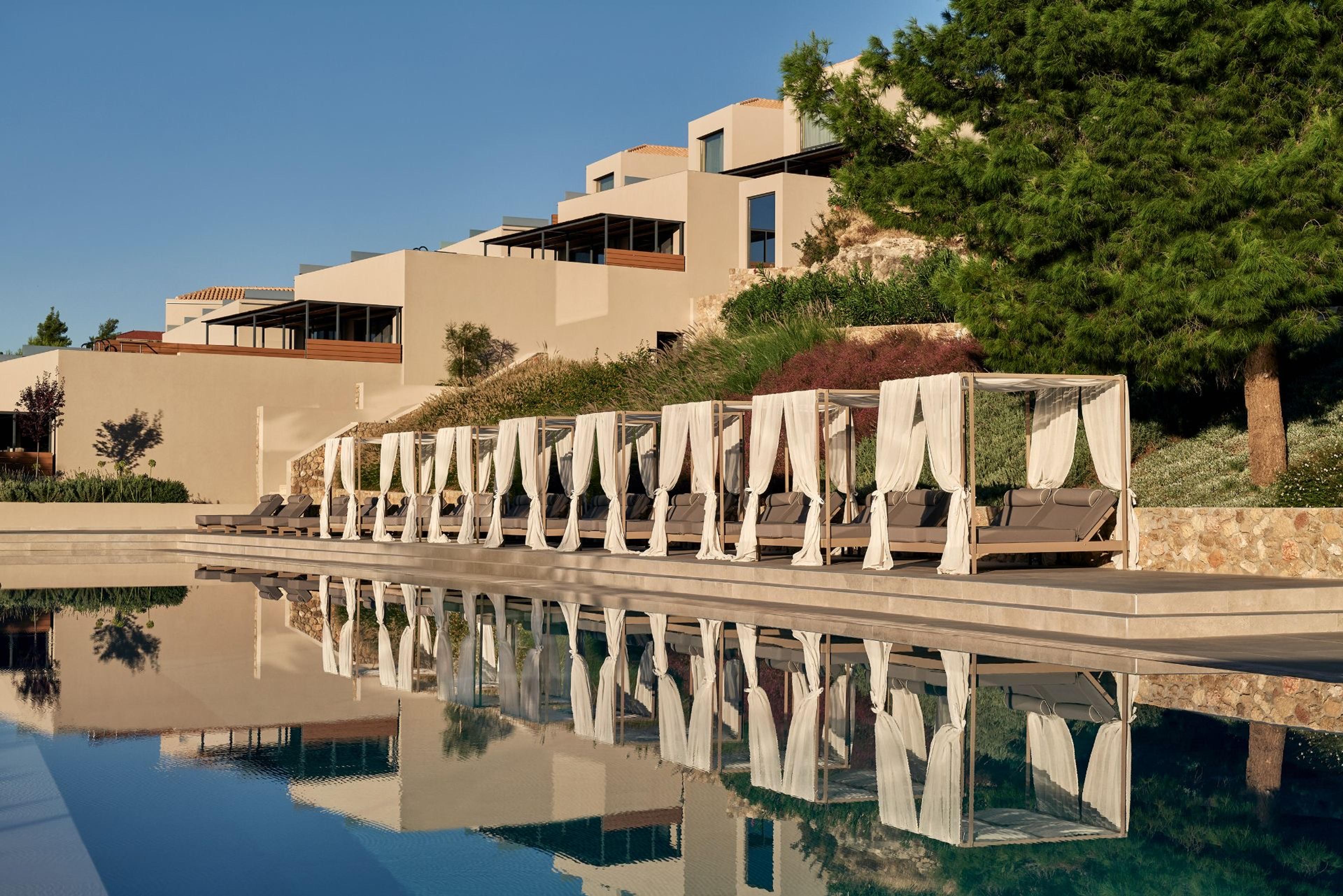 A row of curtained daybeds along an infinity pool, reflecting a tiered hotel building and trees on the hillside behind.