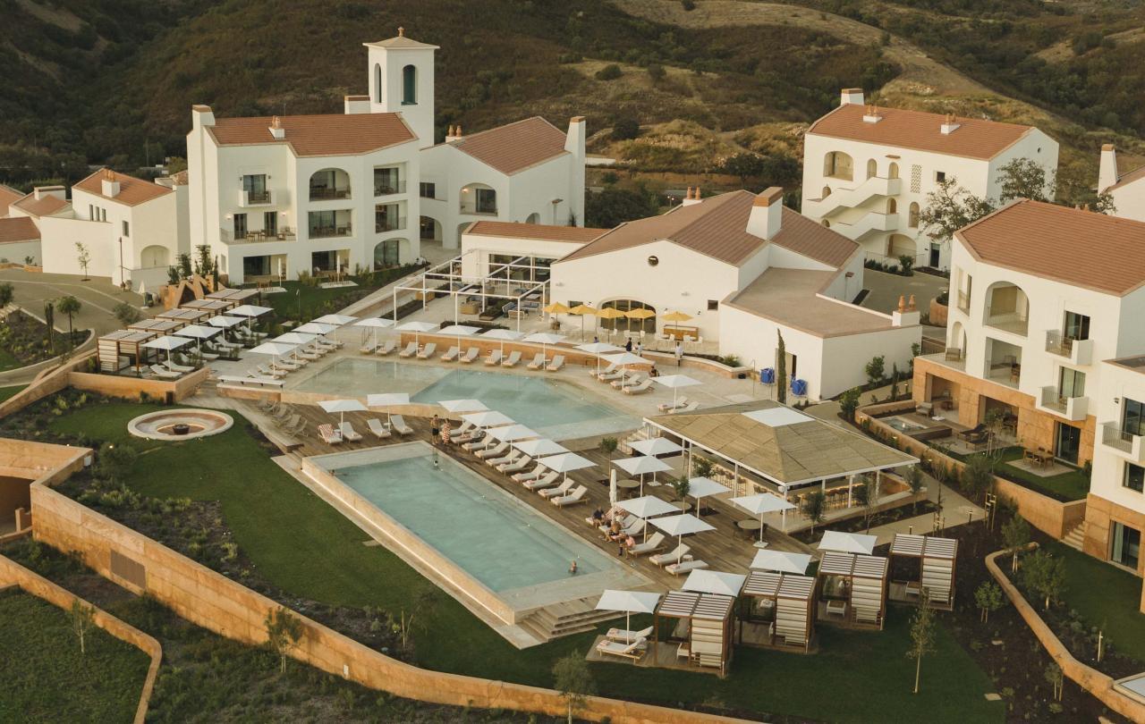 An aerial view at dusk of a resort's pool terrace, with sun loungers, cabanas and whitewashed buildings with terracotta roofs set against a hillside.