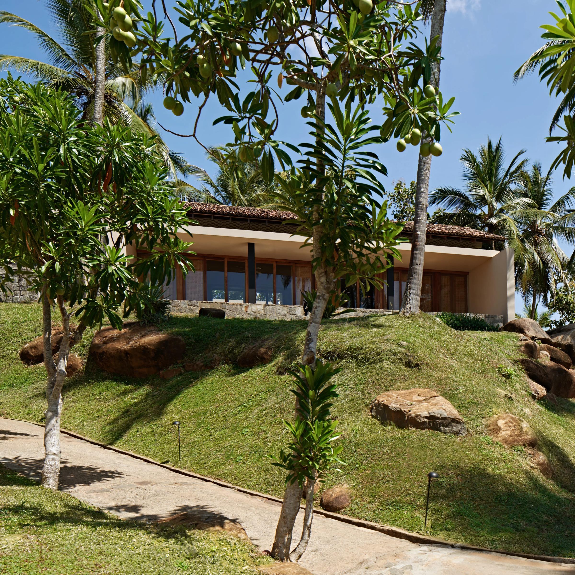 A low villa with a tiled roof seen through mango and palm trees, reached by a stone path across a grassy slope.
