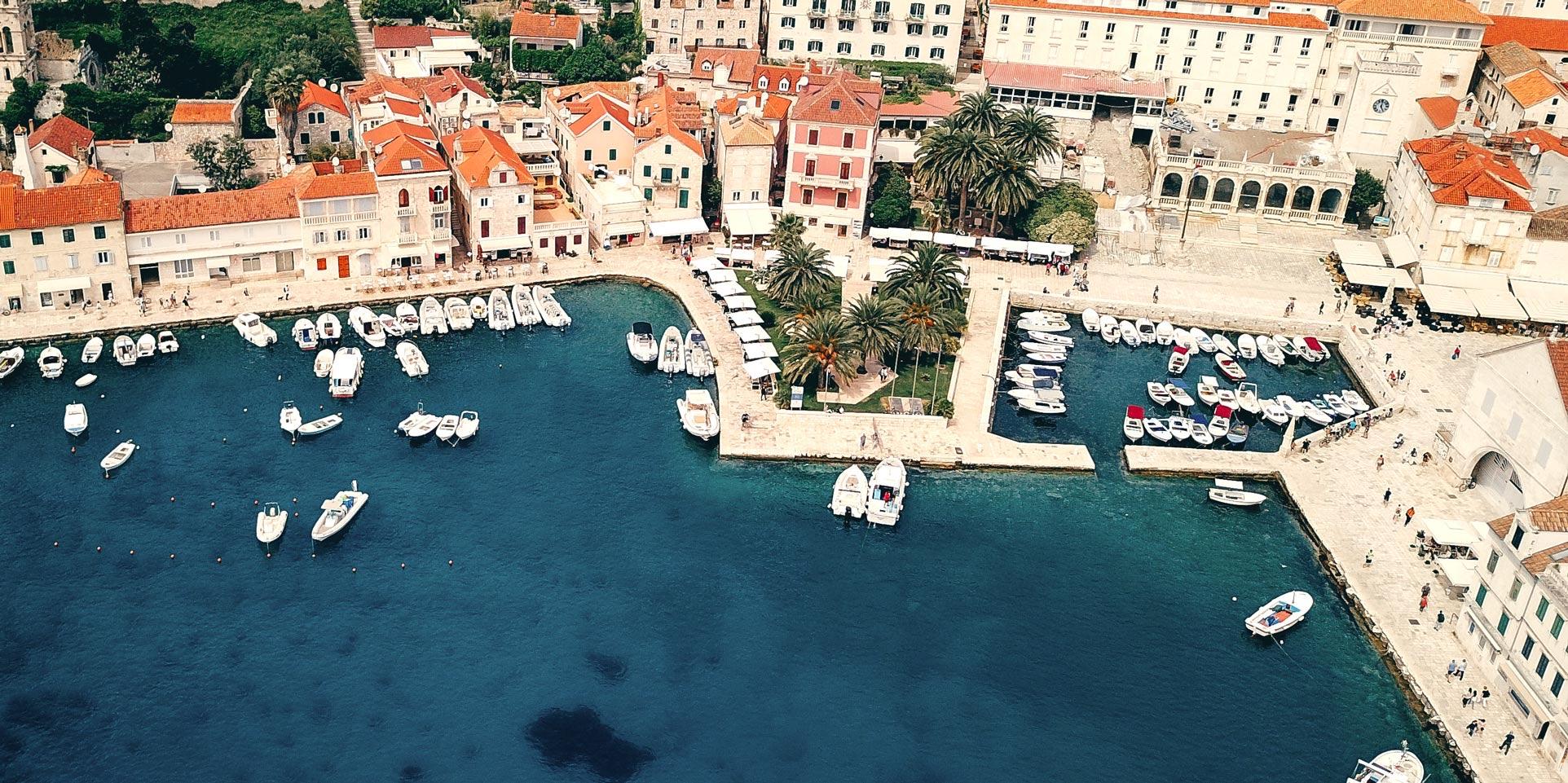 An aerial view of a stone harbour town, with red tile roofs, white motor boats moored along the quays, palm trees around a small square, and clear turquoise water.