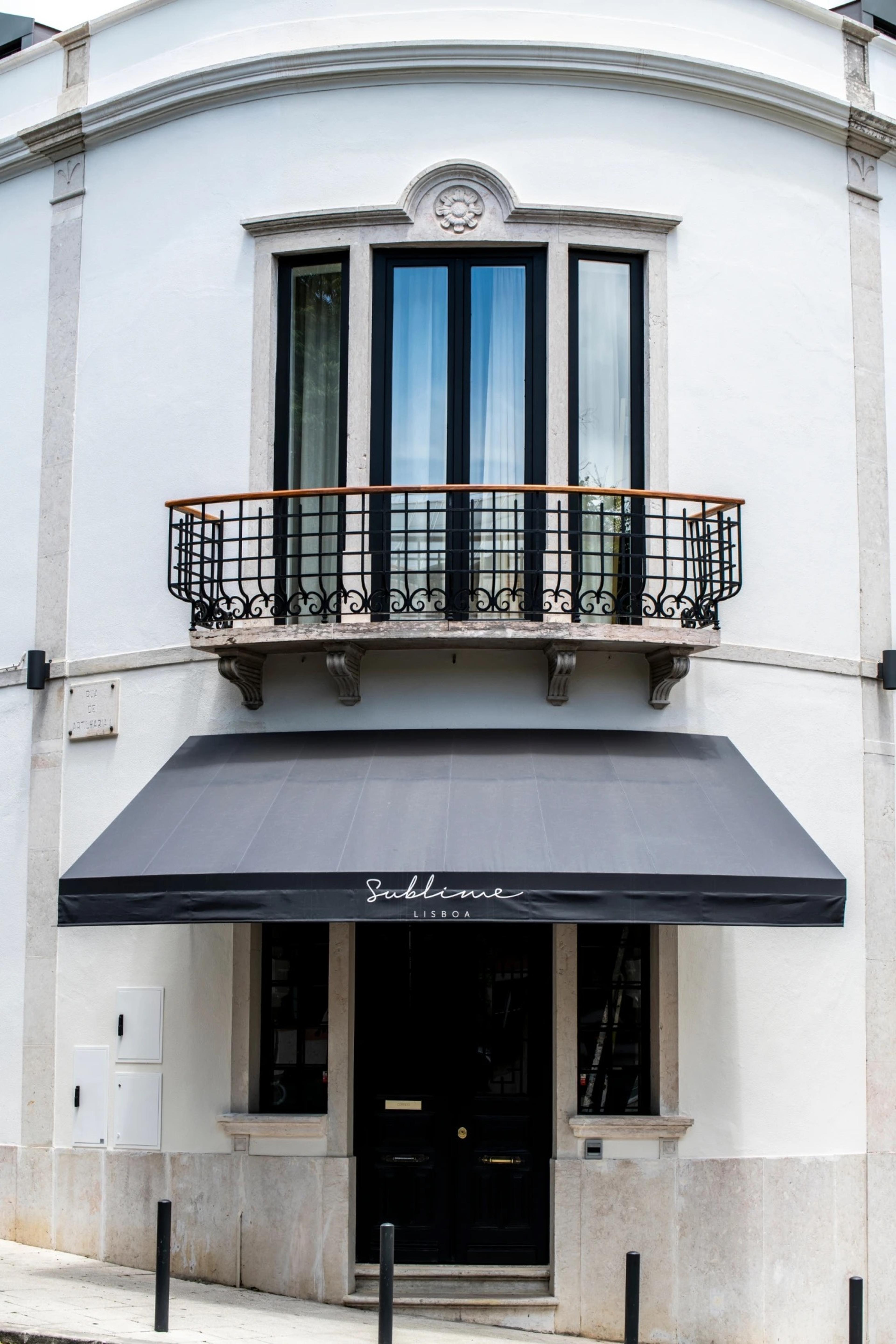 The front of a white corner building with a curved top, a dark wrought-iron balcony, and a grey awning over a black door, the awning reading Sublime Lisboa.