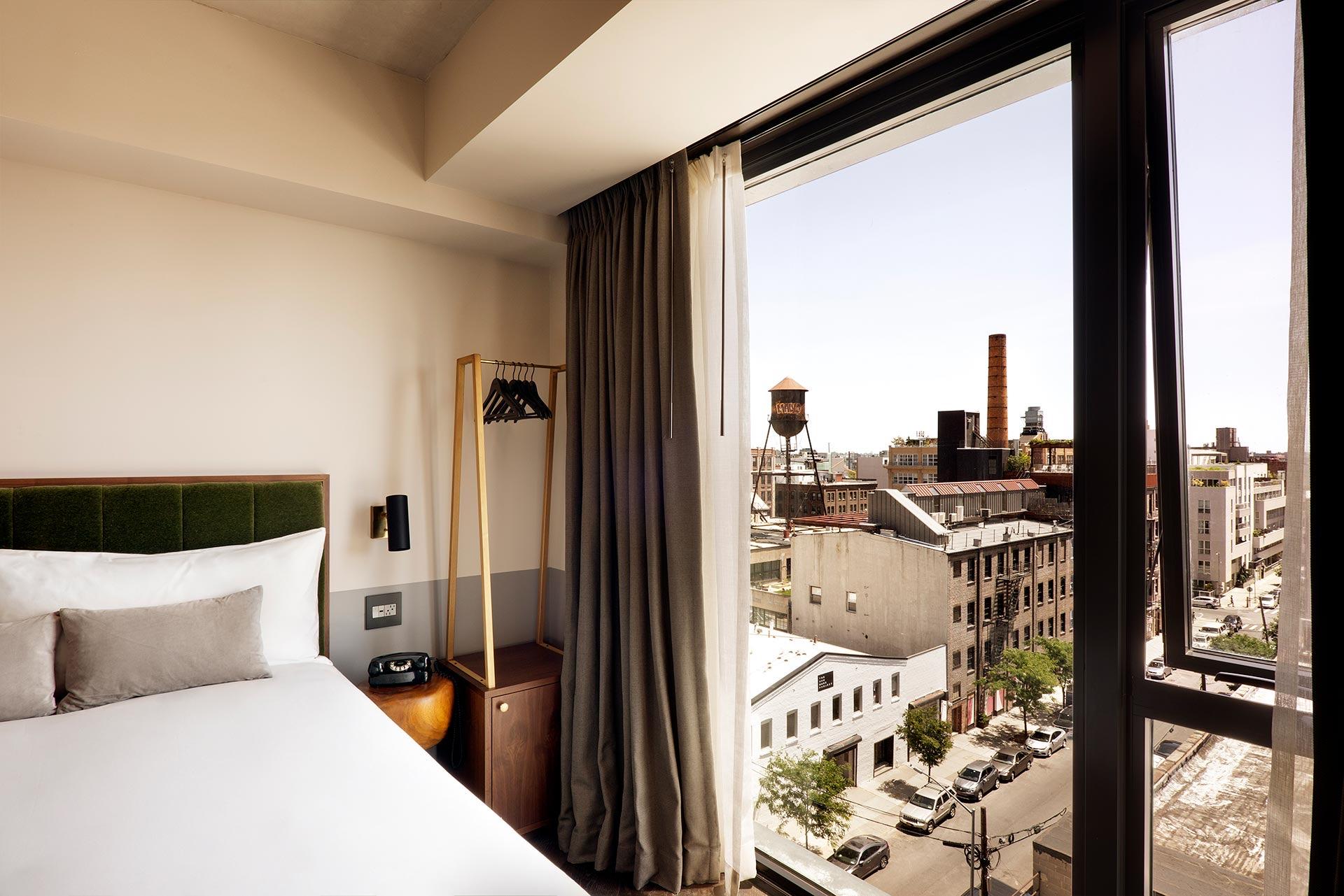 a bed and clothing rail beside an open window looking over a water tower, a smokestack and a street lined with parked cars