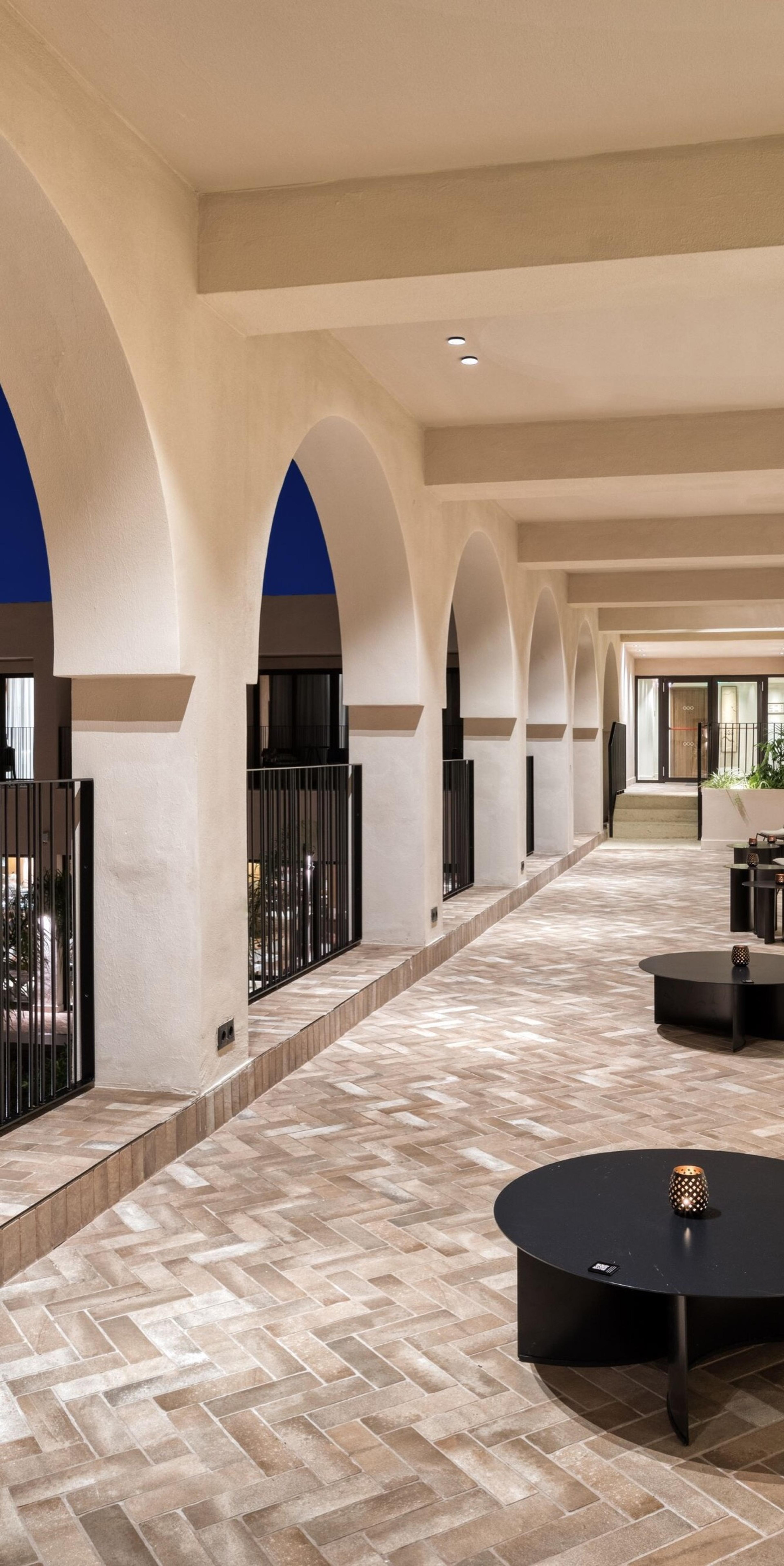 A covered stone arcade with a row of arches opening onto a dark blue dusk sky, with low round tables set with candles down the corridor.