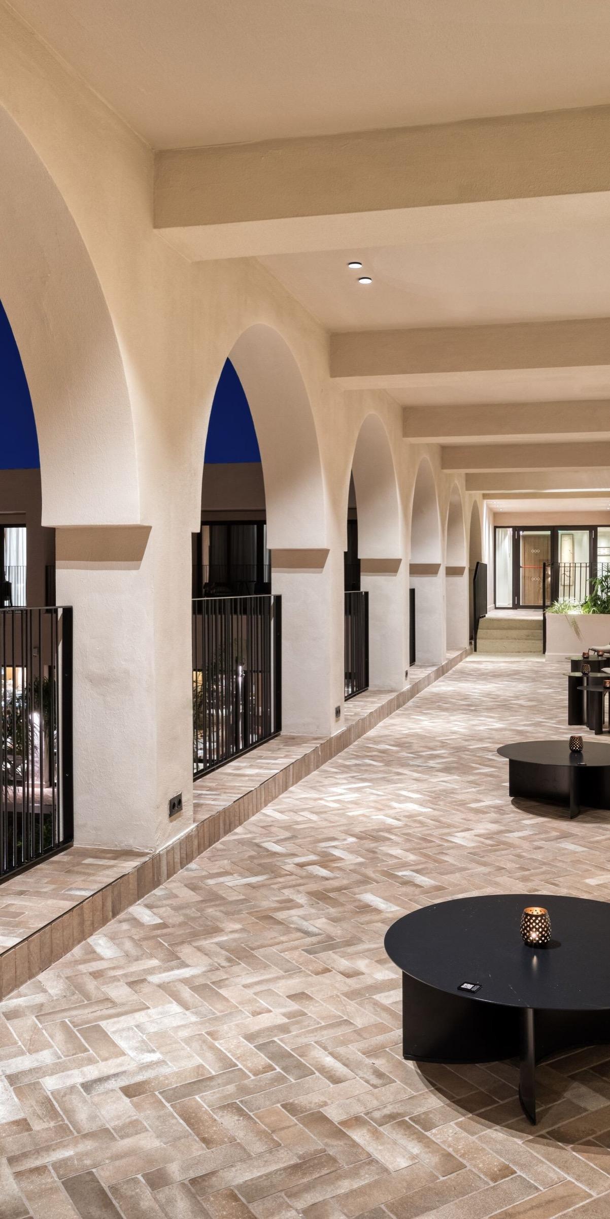 A covered stone arcade with a row of arches opening onto a dark blue dusk sky, with low round tables set with candles down the corridor.