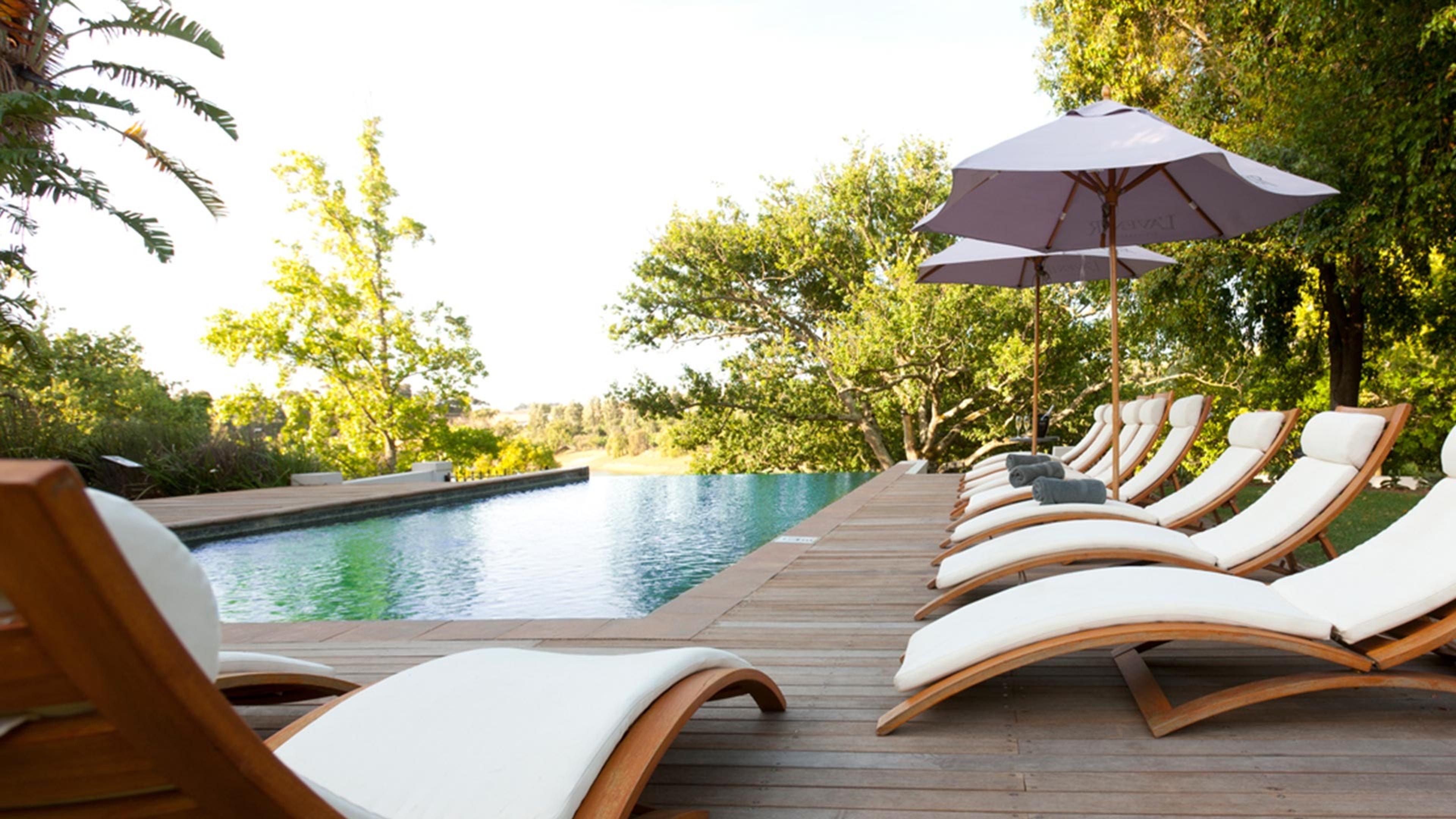 A long pool beside a timber deck, with white-cushioned loungers and grey parasols, and trees behind.