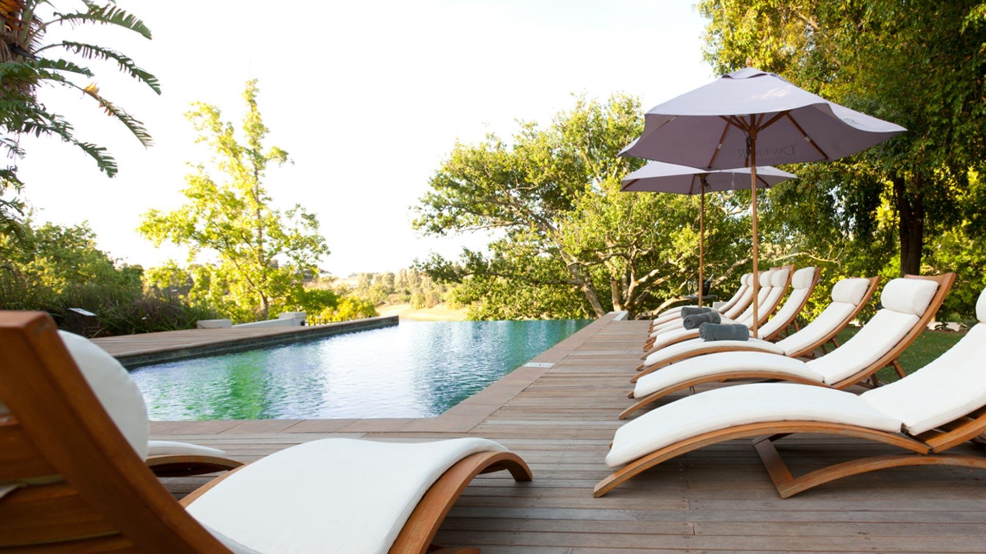 A long pool beside a timber deck, with white-cushioned loungers and grey parasols, and trees behind.