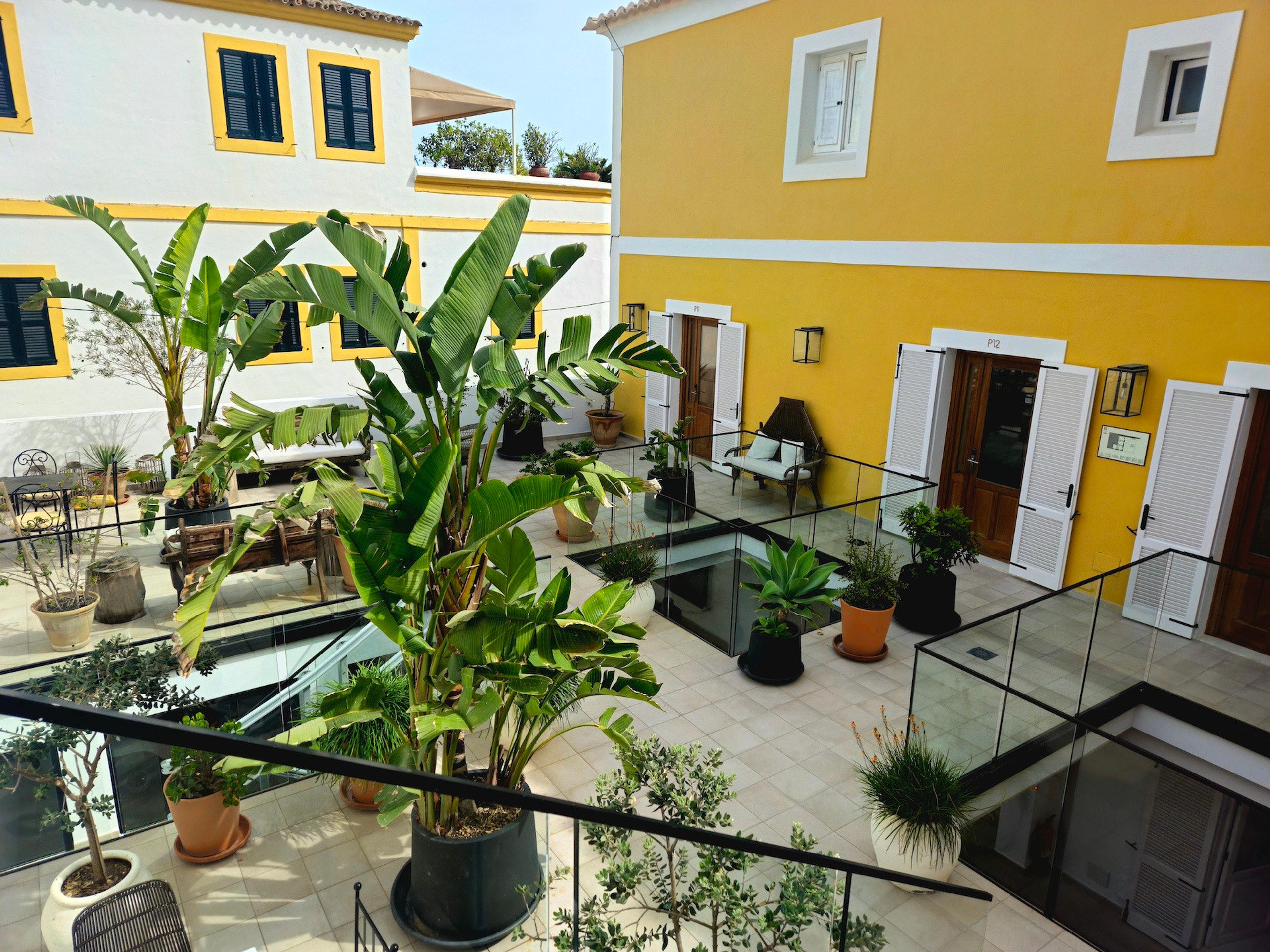A courtyard seen from an upper walkway, with banana palms and potted plants, a yellow-walled building on the right, and glass balustrades.